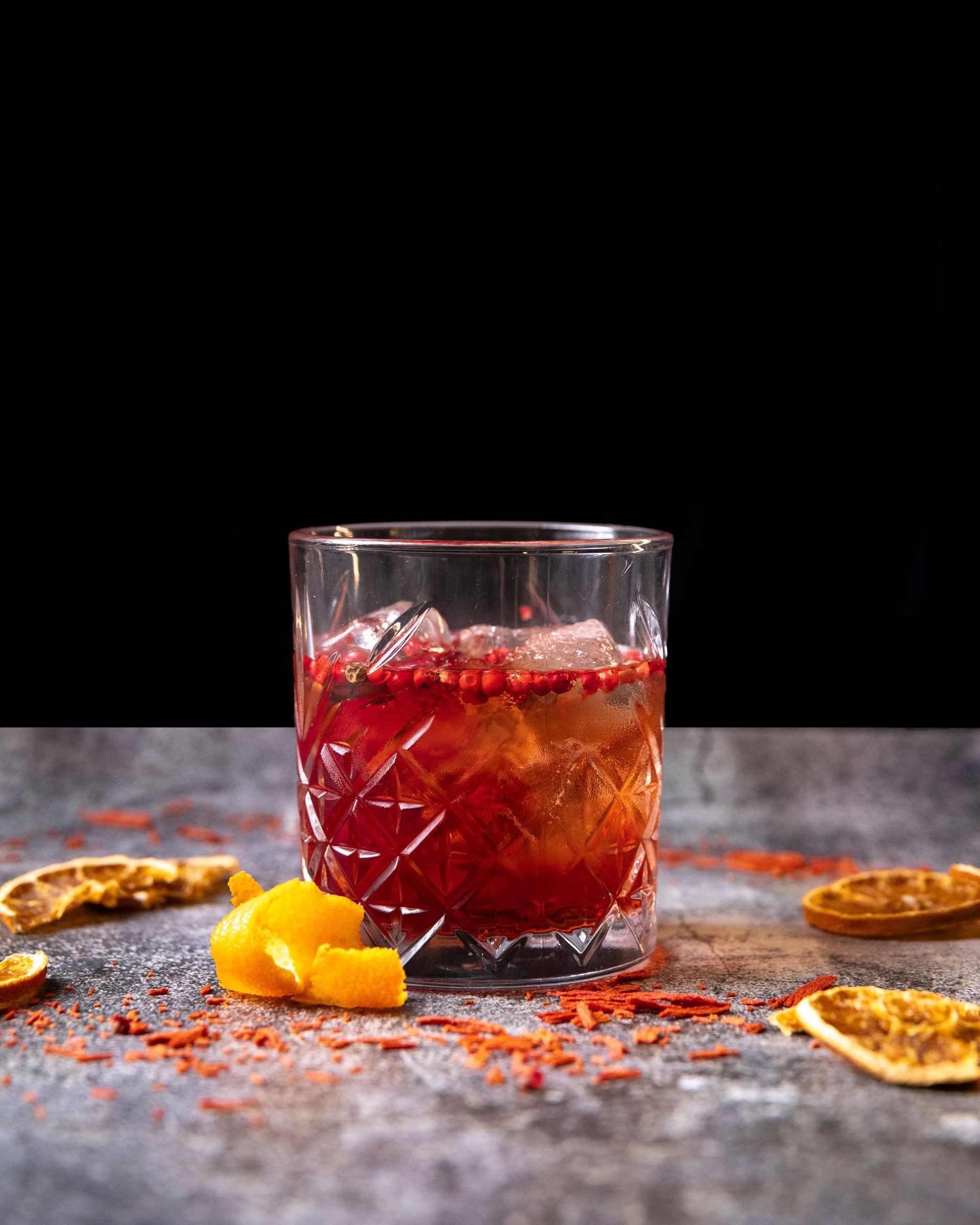 Un cocktail vivace servito in un bicchiere di cristallo con ghiaccio, guarnito con scorza d'arancia e grani di pepe rosso, su una superficie grigia testurizzata con fette d'arancia essiccate.