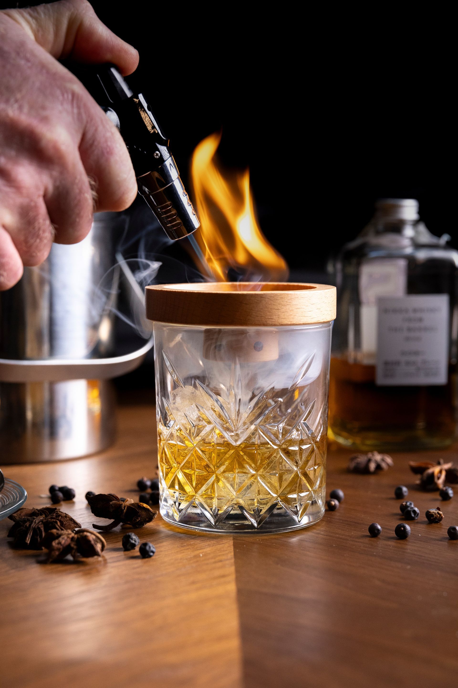Una mano brucia il bordo di un bicchiere da cocktail con una fiamma, infondendo fumo. Anice stellato e grani di pepe sono sparsi su una superficie di legno.