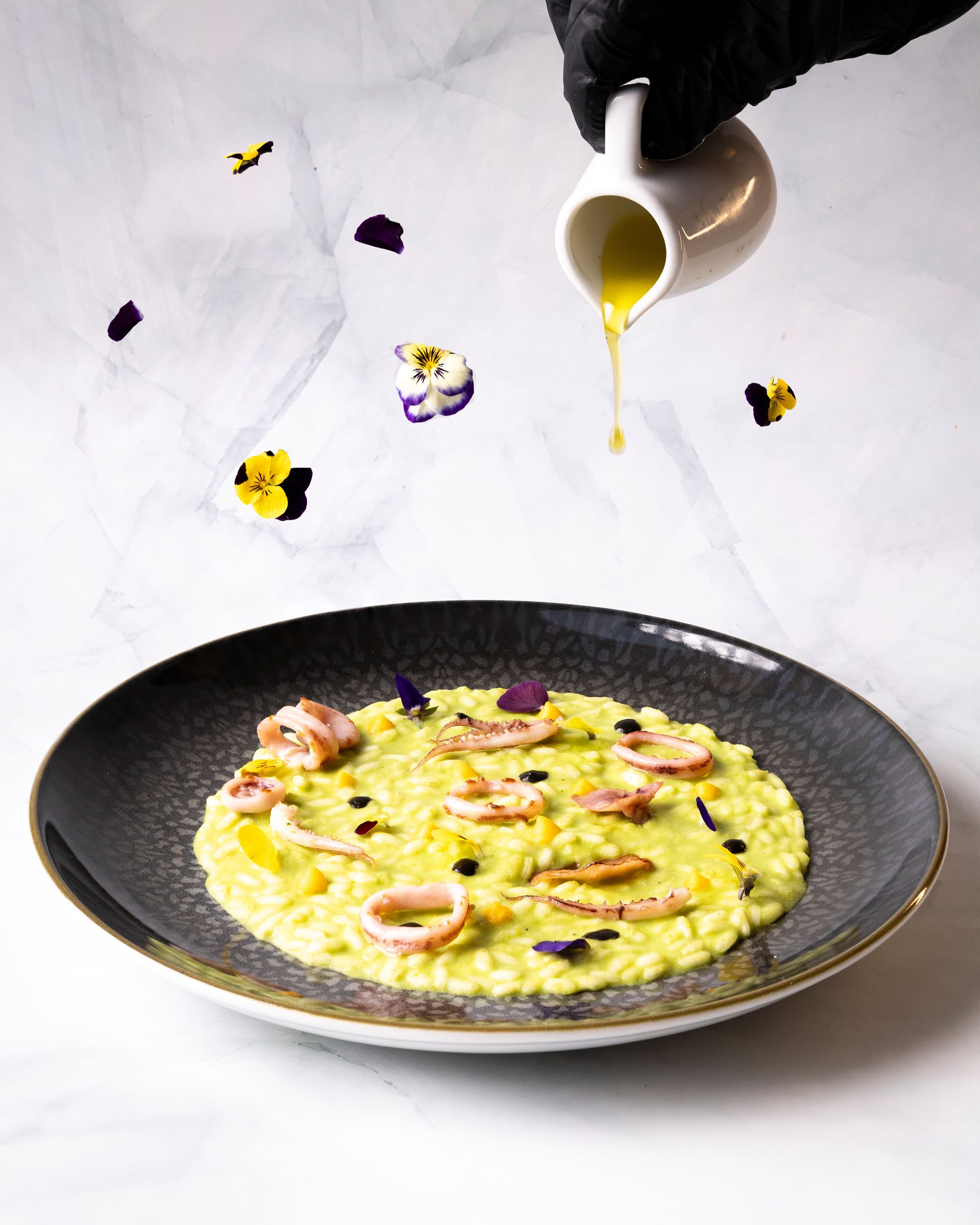 Un piatto di risotto giallo con calamari e salsa servita in una brocca bianca tenuta da una mano guantata; il tutto decorato con fiori commestibili.