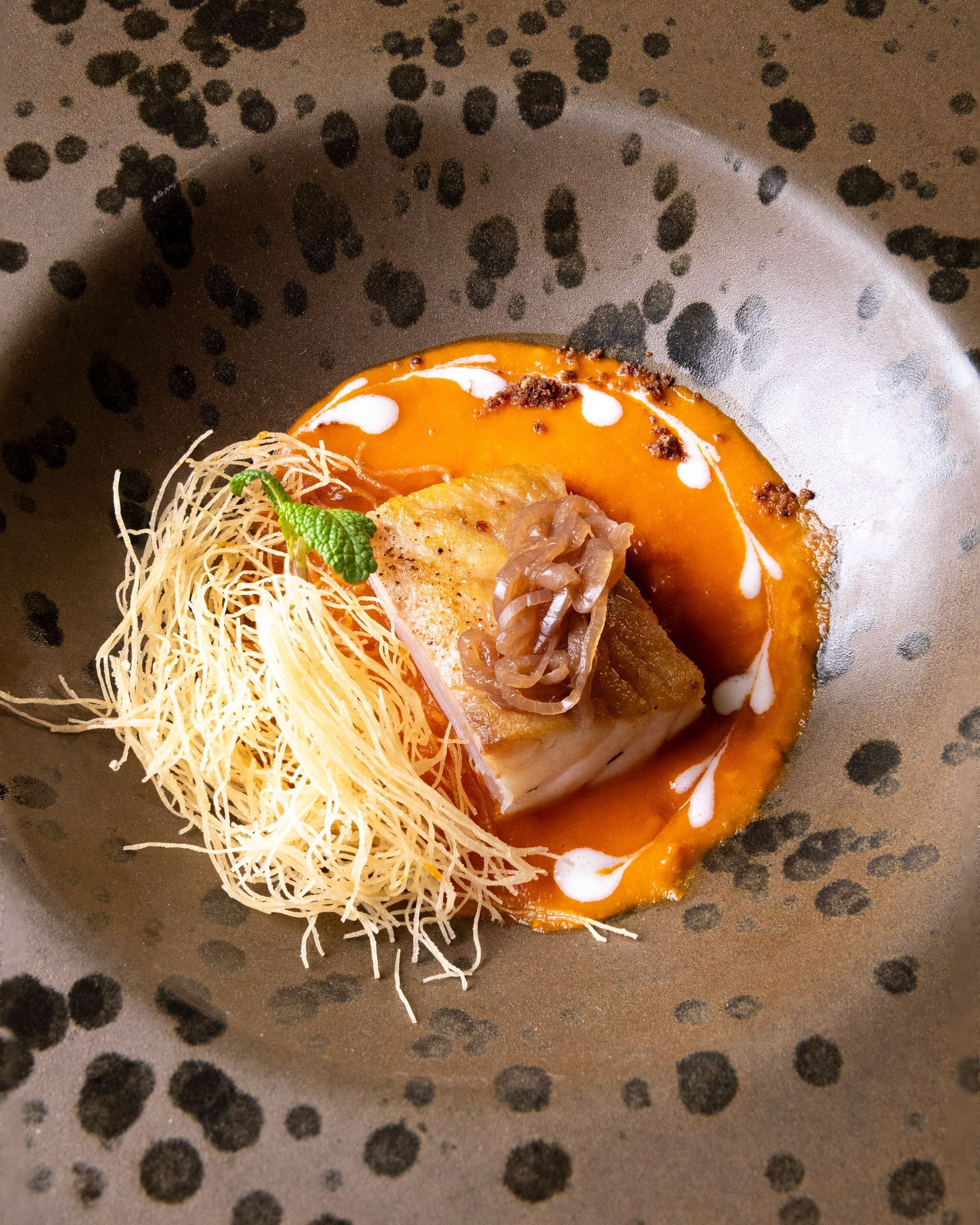 Primo piano di un piatto gourmet: pesce scottato con salsa e nido di noodles croccanti su un piatto decorativo.