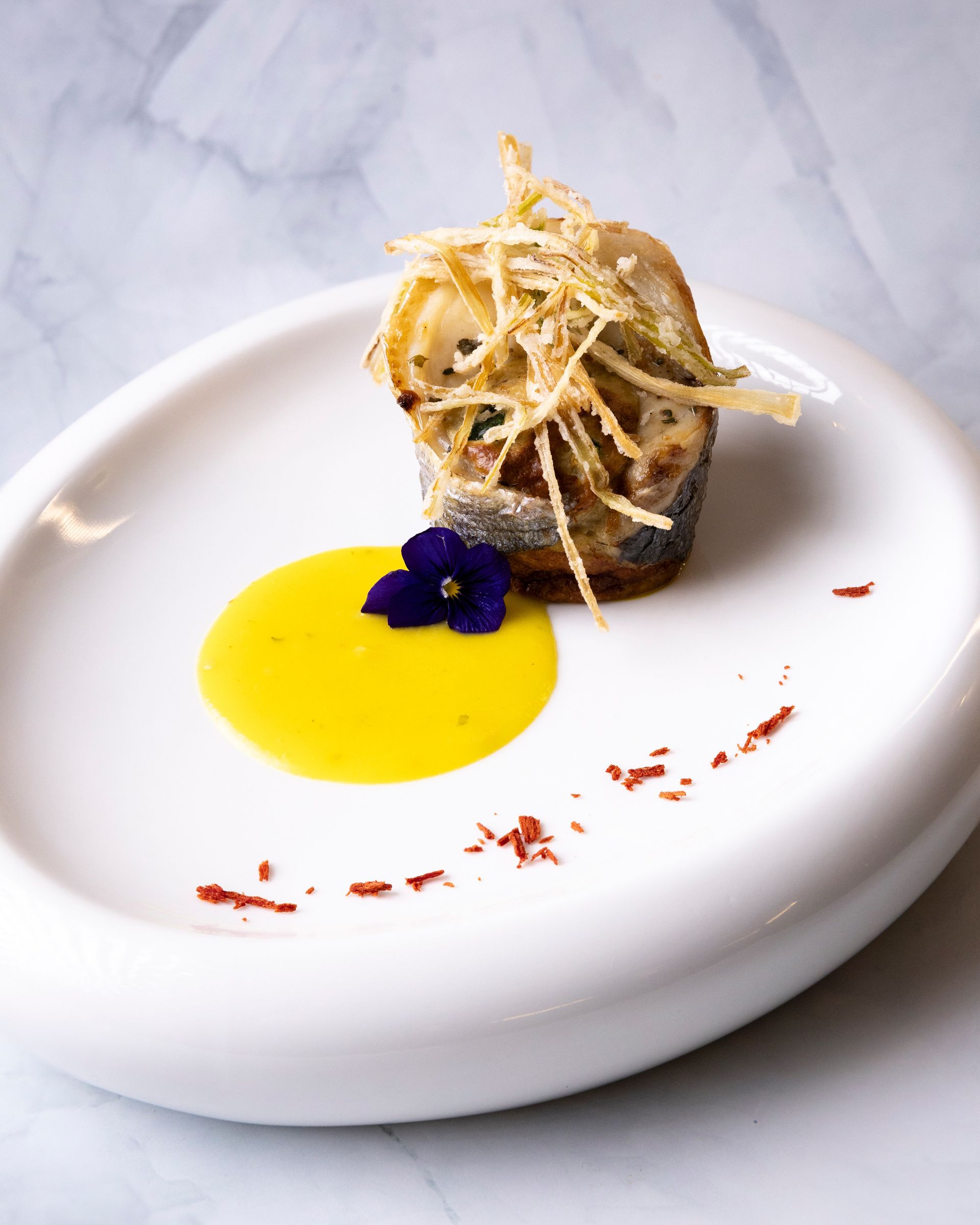 Piatto elegante con pesce, salsa gialla e fiori viola. Il pesce è ricoperto da una croccante copertura, su uno sfondo di marmo.