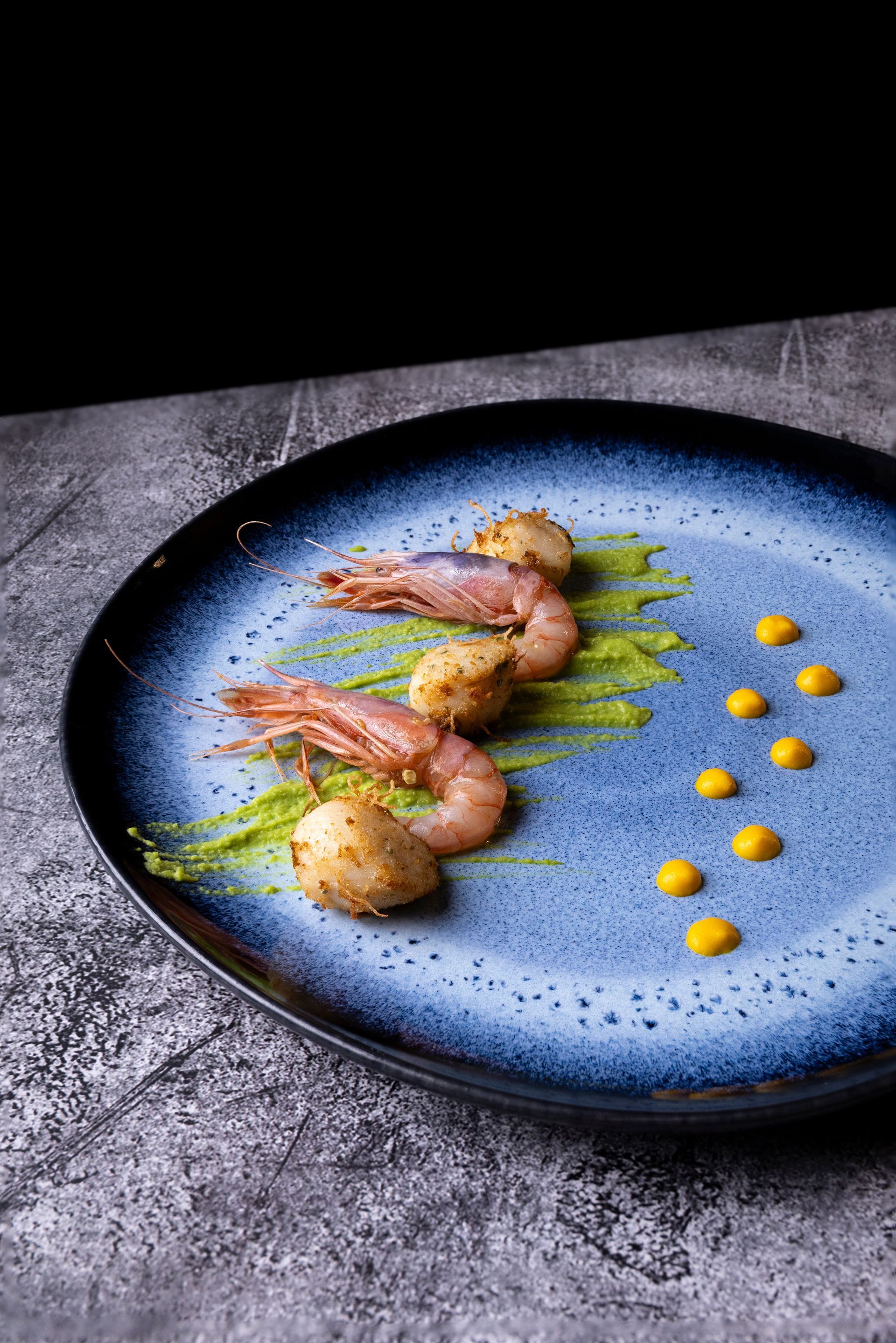 Piatto impiattato con gamberi grigliati e capesante di patate con salse verdi e gialle. Il piatto ha un bordo blu su una superficie grigia.