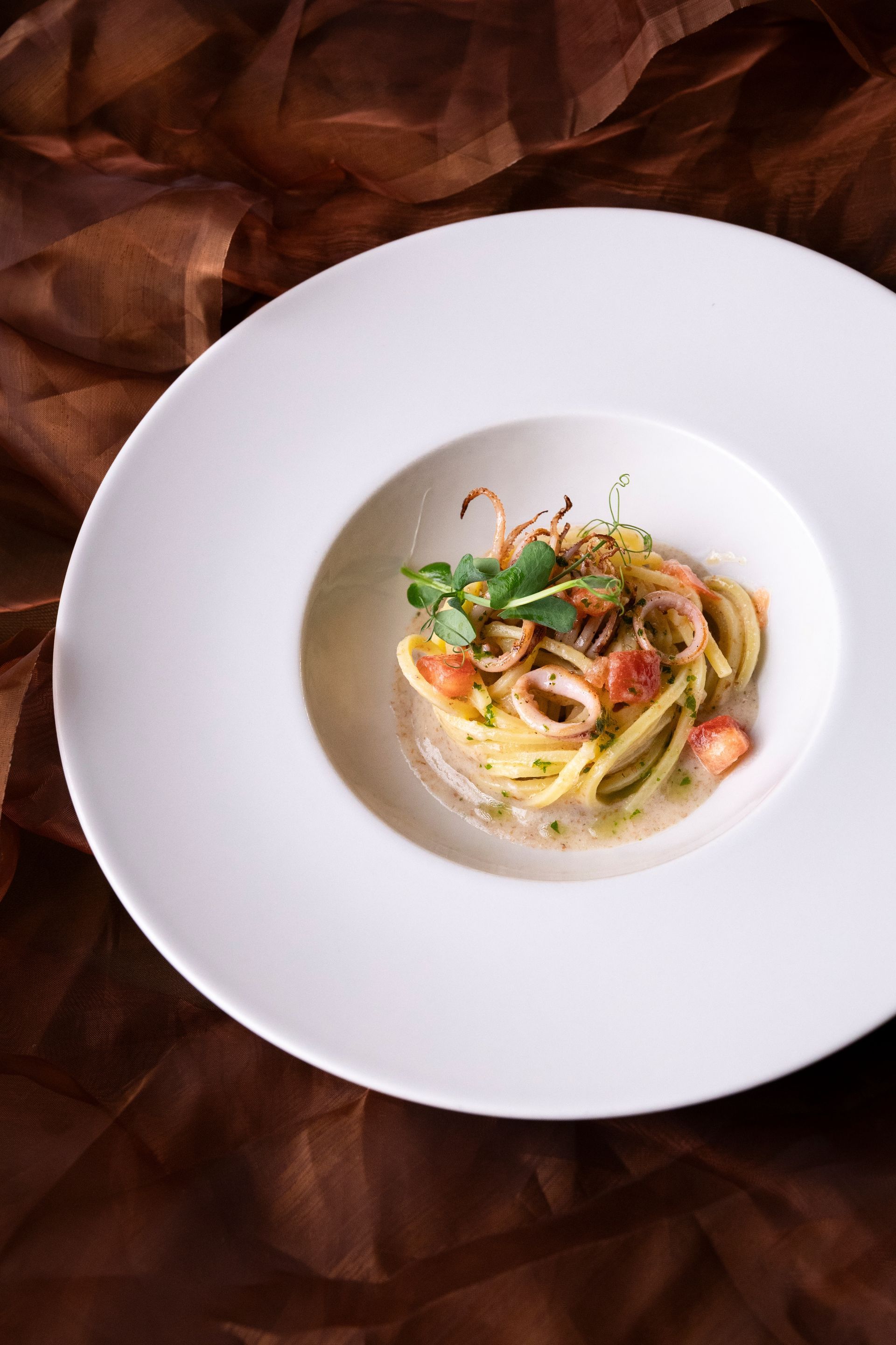 Piatto di pasta con calamari, erbe aromatiche e salsa in una ciotola bianca su un panno marrone.