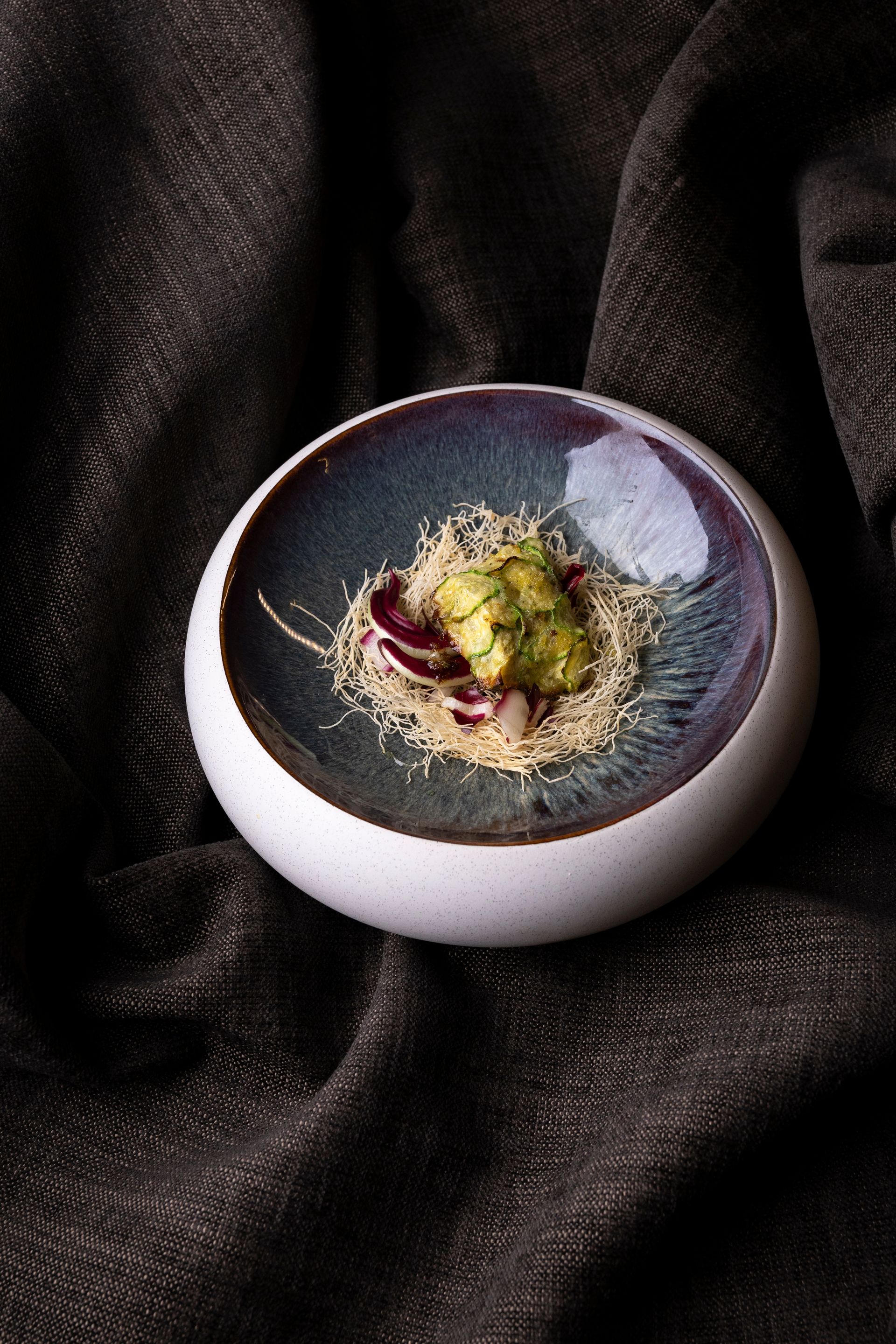 Piccola ciotola con bordo bianco e interno blu-viola, contenente un alimento verde con accenti rossi e semi di sesamo, su una superficie scura e ruvida.