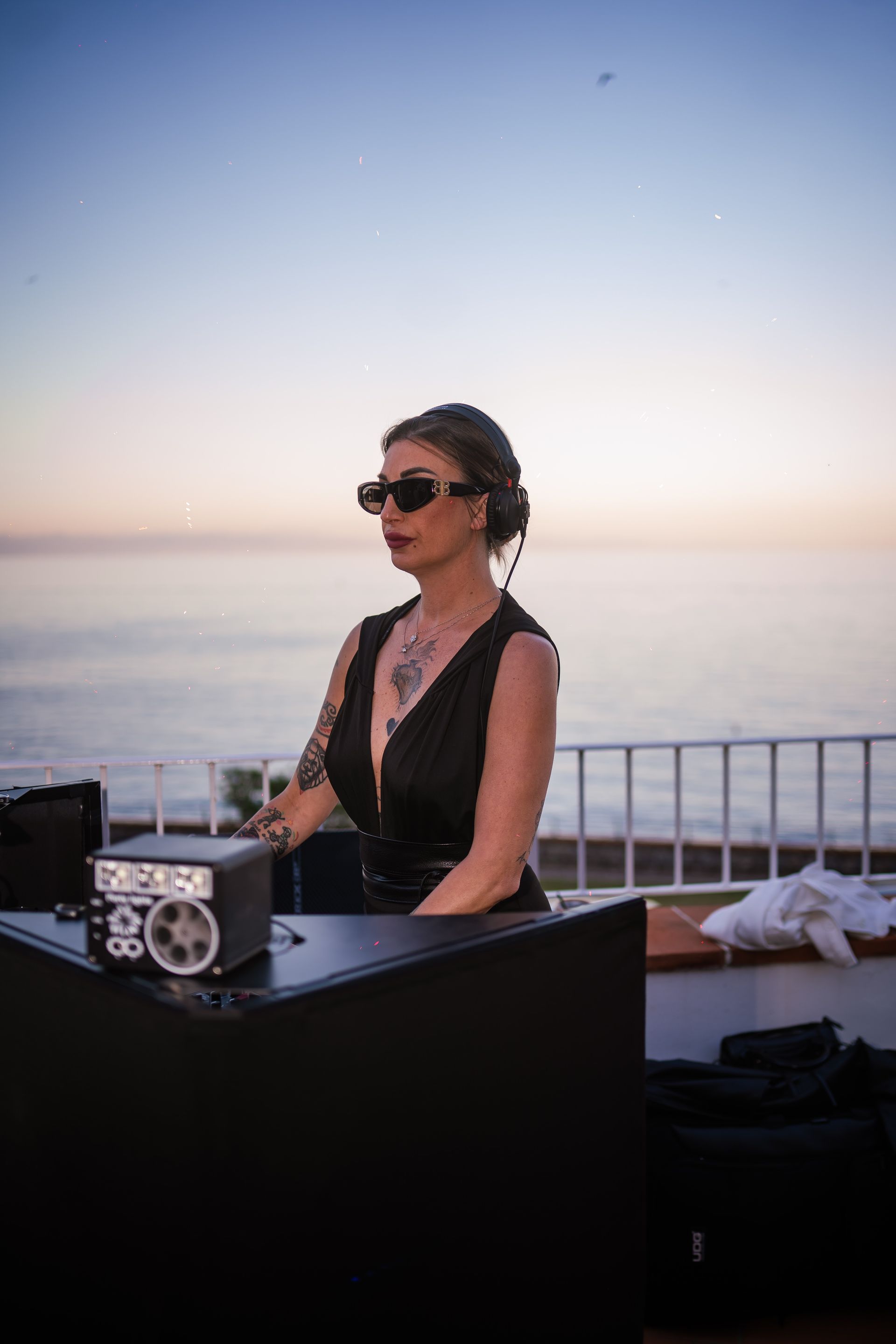 DJ con occhiali da sole, tatuaggi e abito nero, che si esibisce all'aperto al tramonto con l'oceano visibile sullo sfondo.