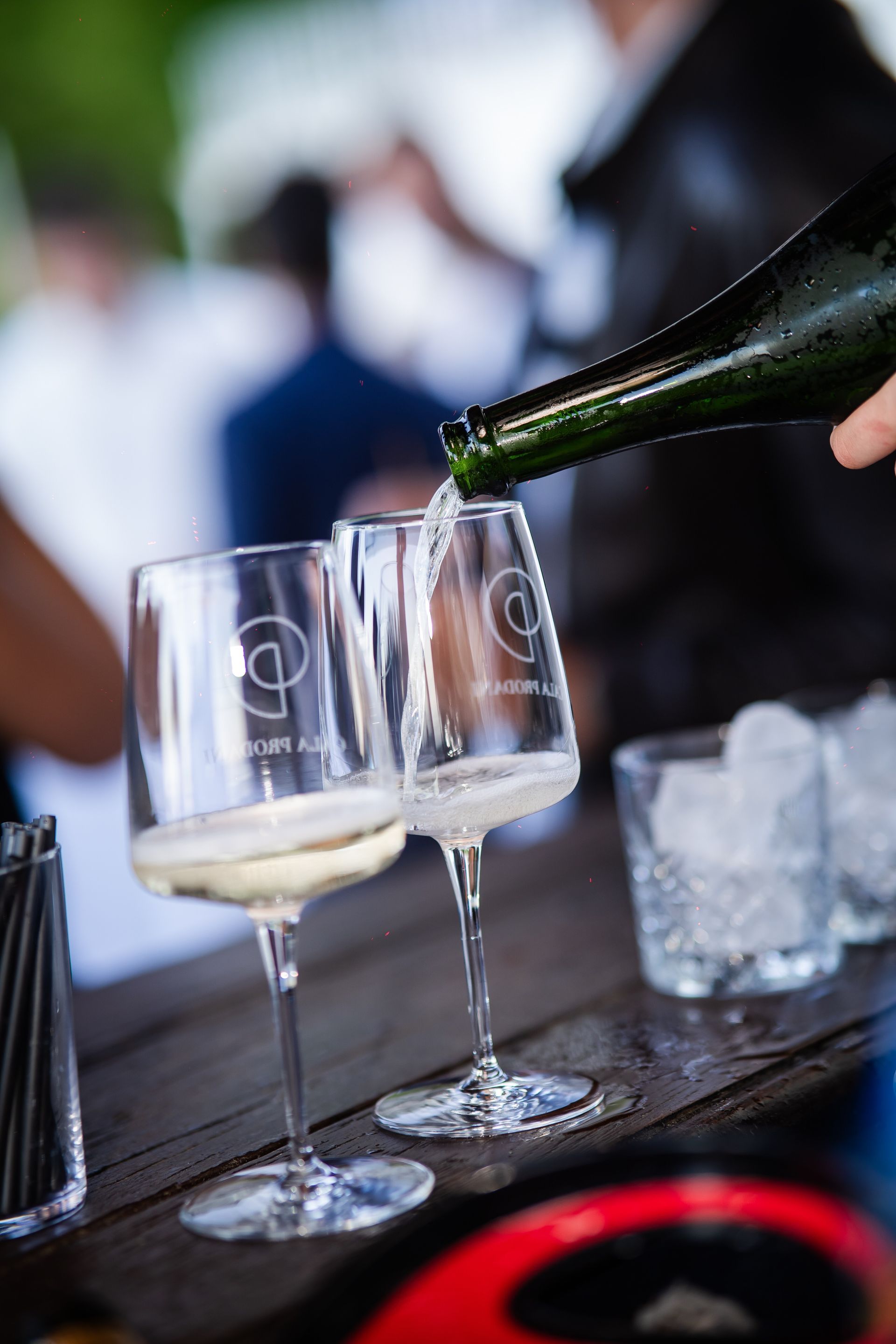 Una persona versa dello spumante in due calici su un bancone di legno. Altre persone sono sullo sfondo sfocato.