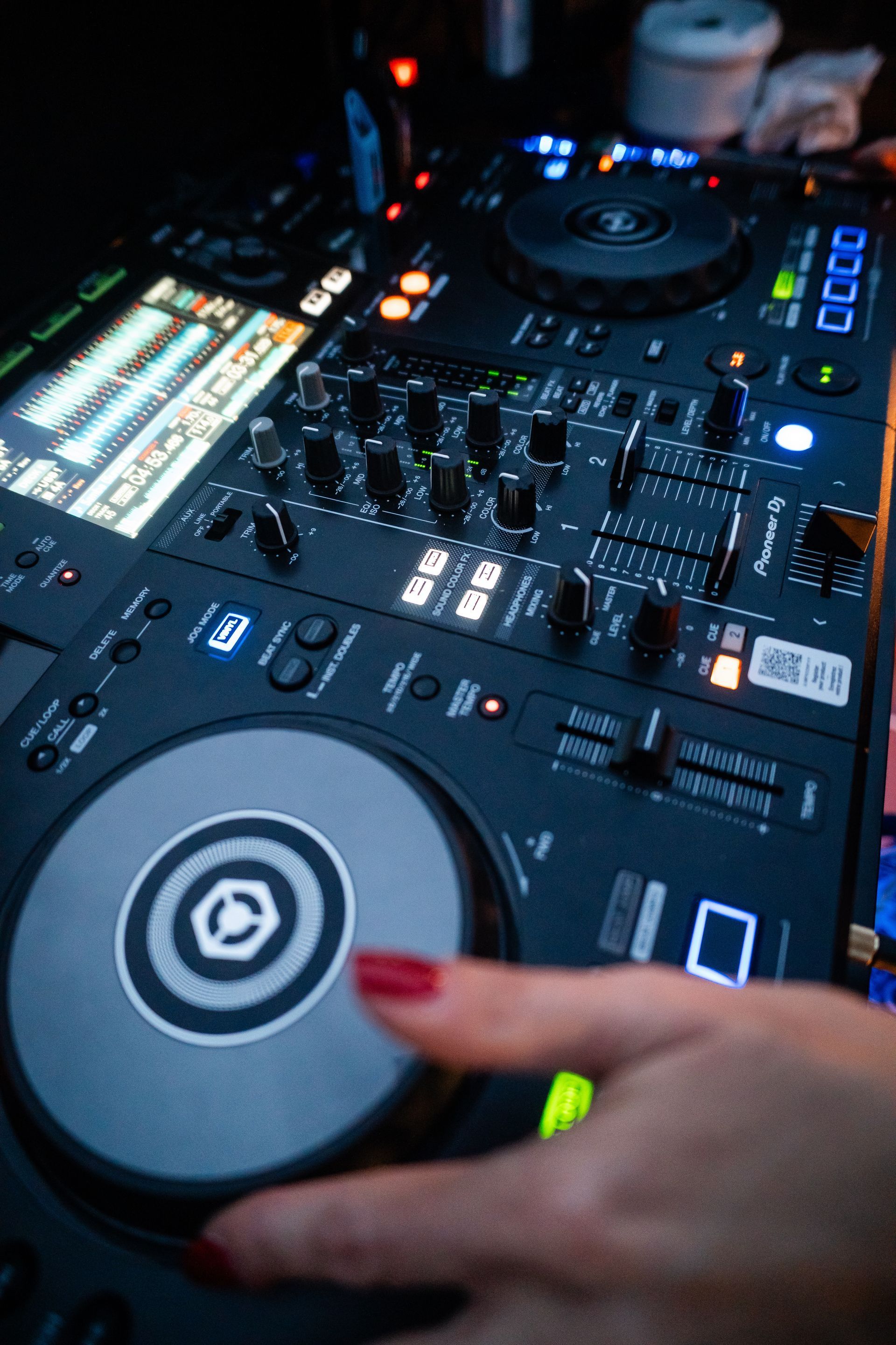 Una mano con le unghie rosse che fa girare un giradischi su un mixer da DJ. Il mixer è nero e presenta vari pulsanti e controlli.