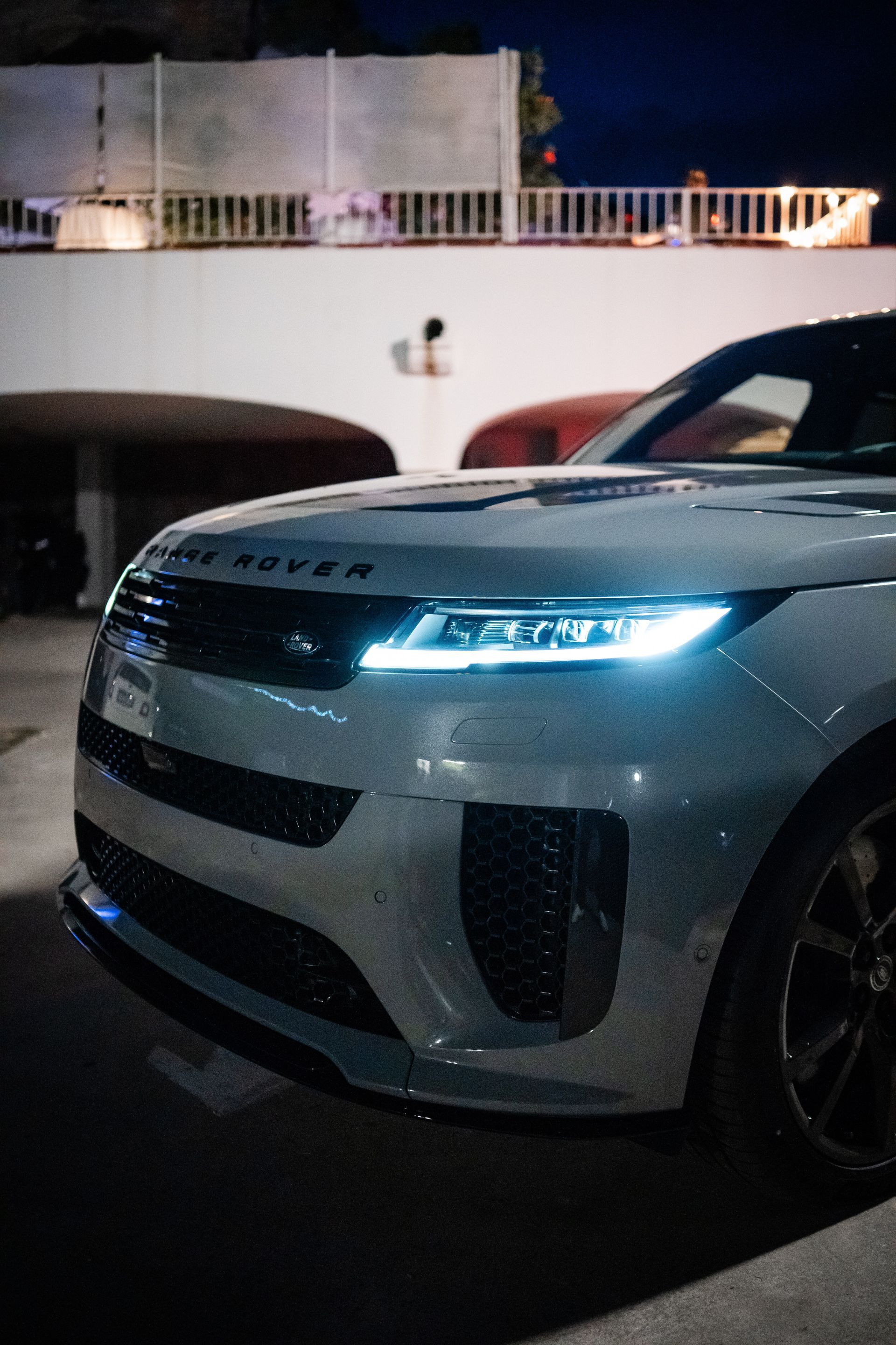 Un SUV Range Rover argentato parcheggiato all'aperto di notte con i fari accesi. L'ambientazione sembra essere un'area urbana.
