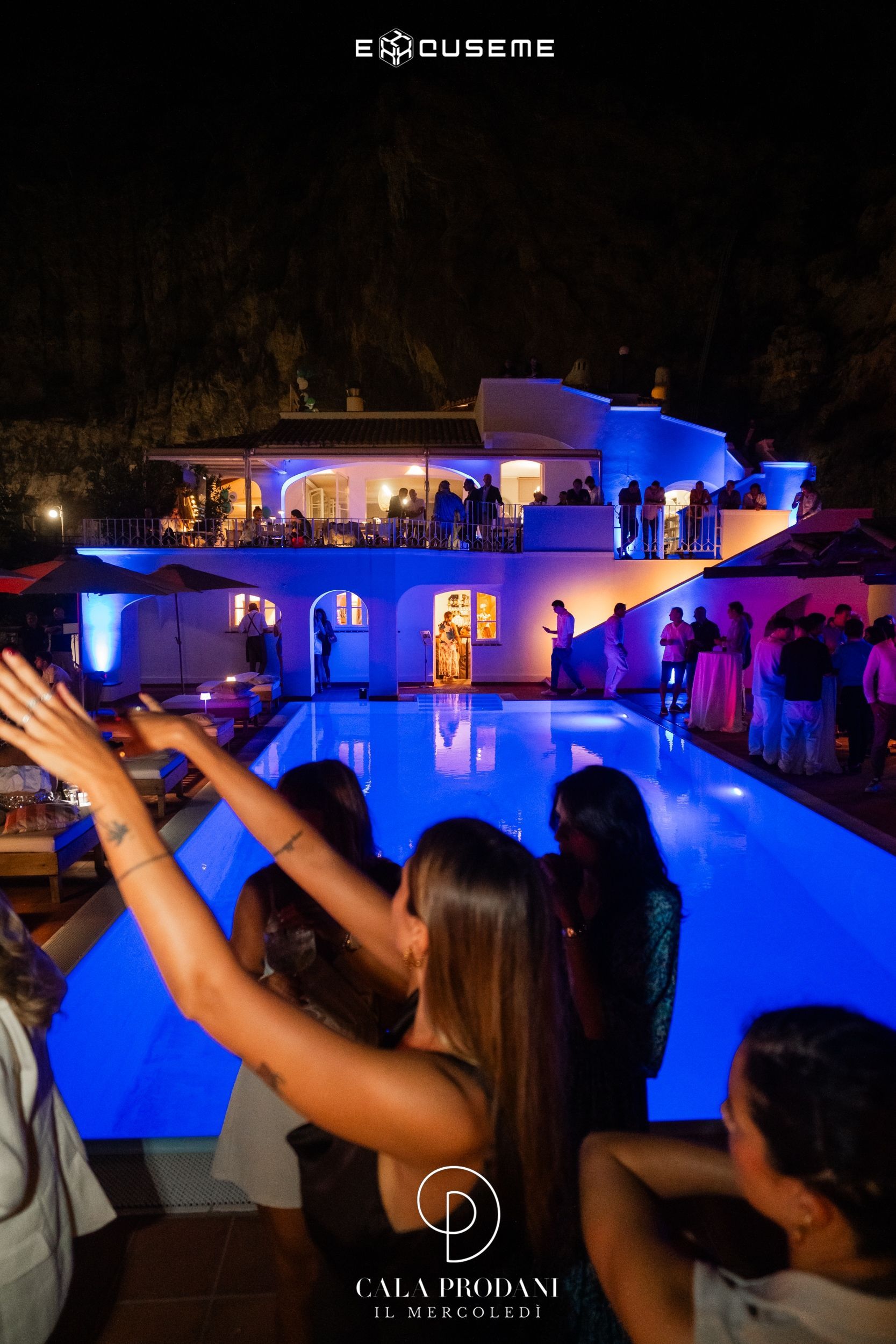 Persone che ballano di notte a bordo piscina in un locale all'aperto con un edificio bianco e luci blu.