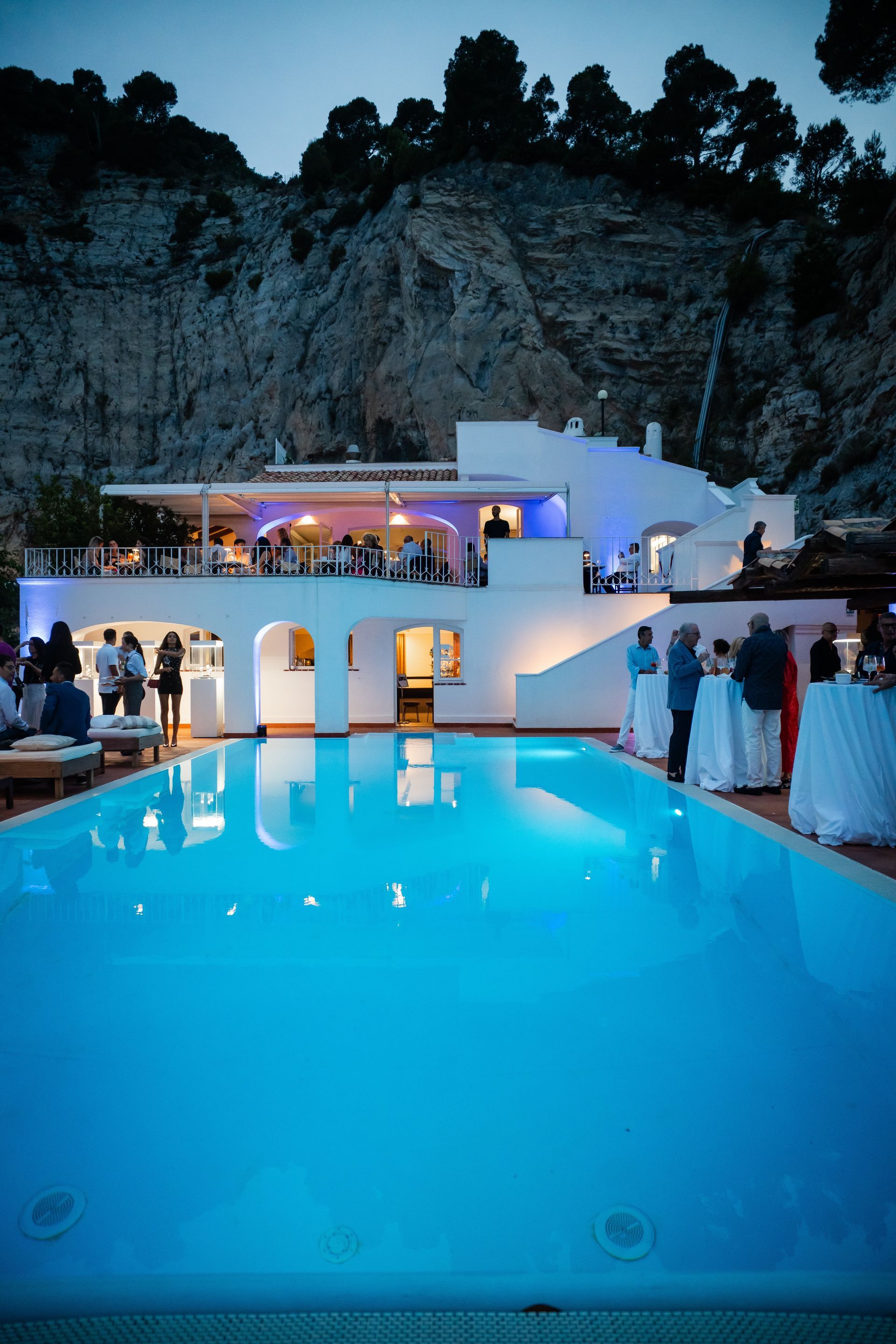 Un edificio bianco con una piscina illuminata di blu, adagiato su una scogliera. La gente si riunisce per un evento serale, con luci soffuse.