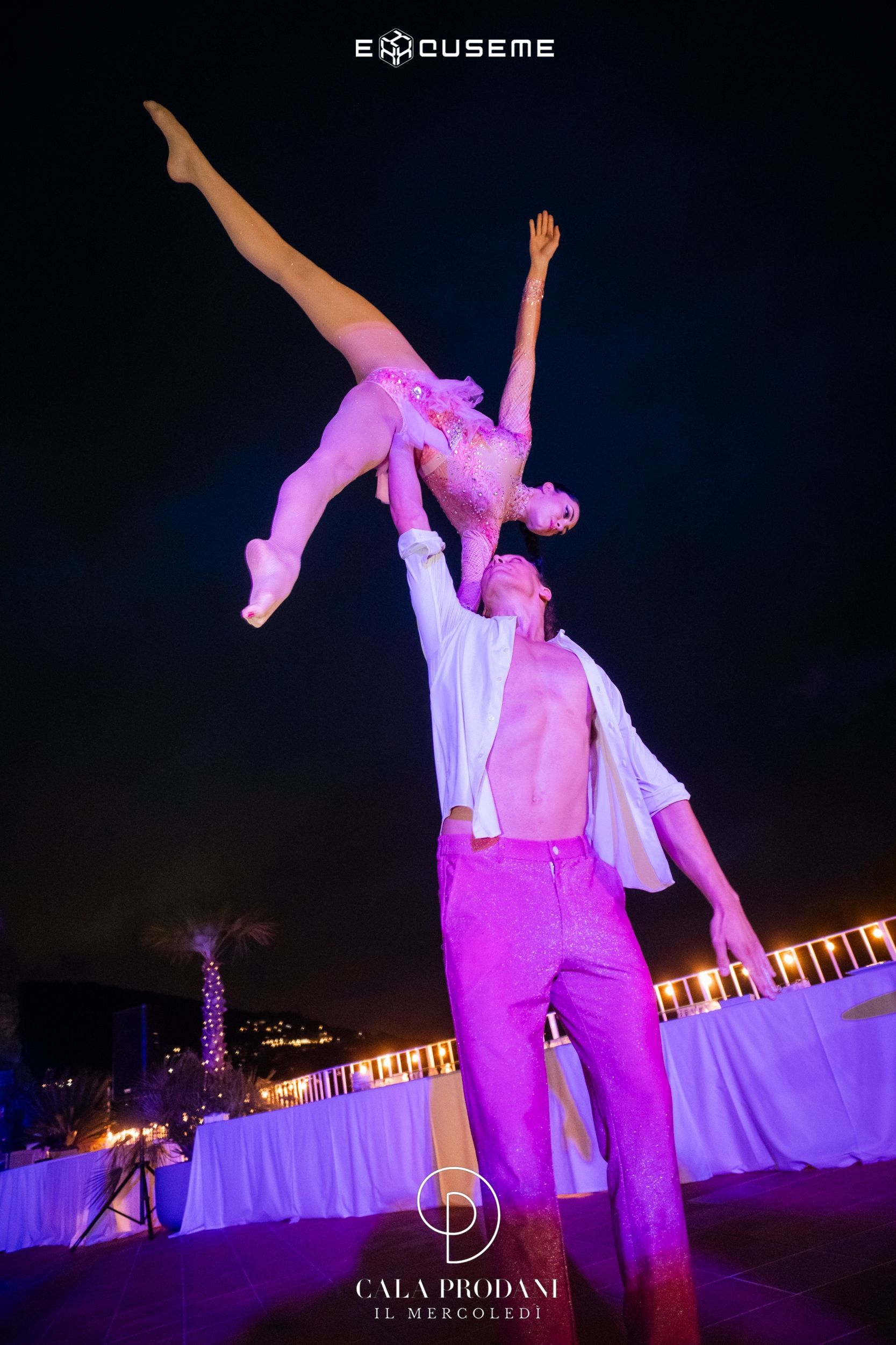 Duo acrobatico che si esibisce di notte. L'uomo tiene in alto la donna, entrambi in scintillanti abiti rosa, con le braccia tese.