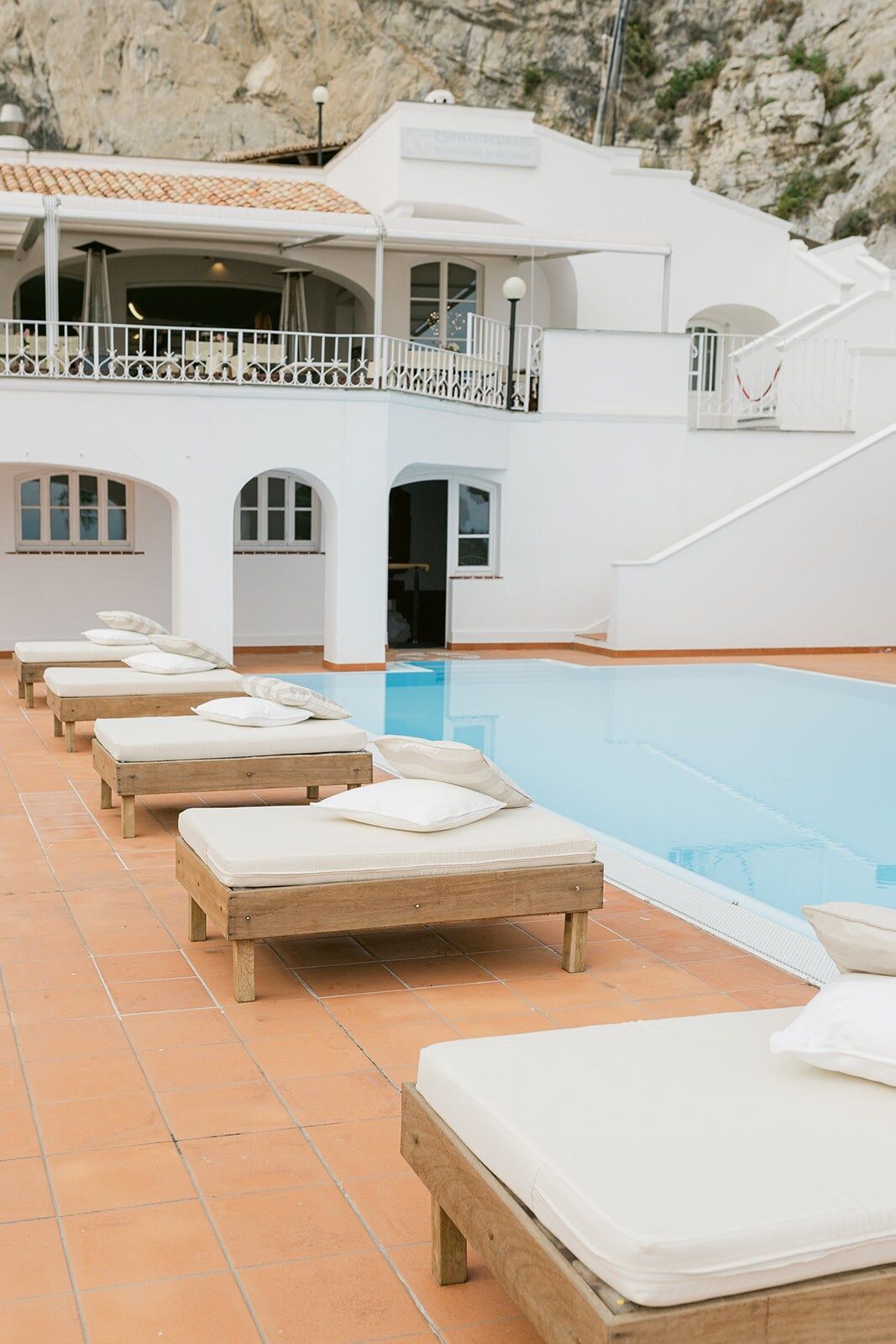 Una scena a bordo piscina con lettini bianchi e una piscina blu di fronte a un edificio bianco; sullo sfondo è visibile una scogliera rocciosa.