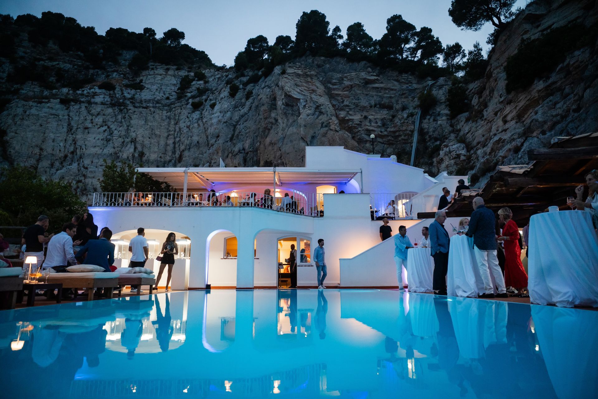 Evento notturno in un edificio bianco con piscina, adagiato su una scogliera. La gente si mescola vicino alla piscina, che riflette le luci dell'edificio.