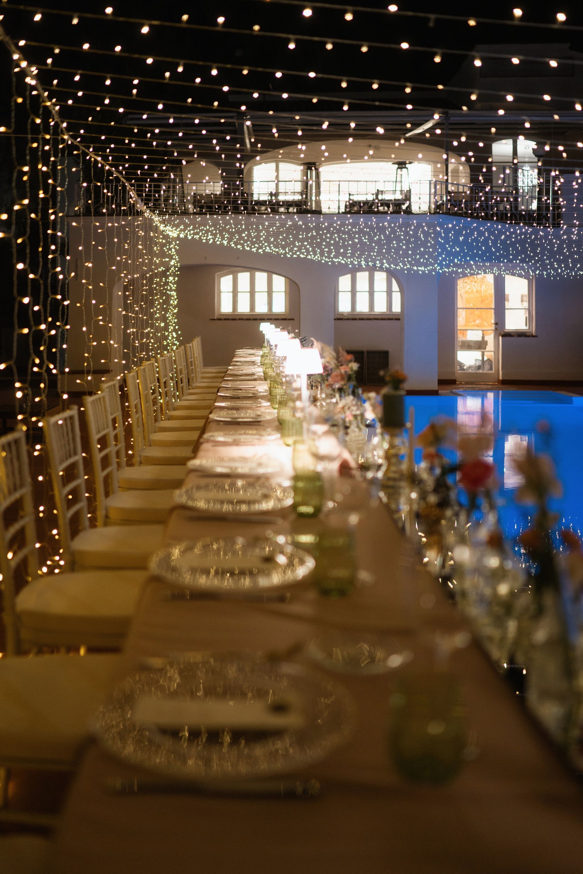 Un lungo tavolo da pranzo apparecchiato per un evento formale. Luci a ghirlanda in alto; sullo sfondo si vede una piscina.