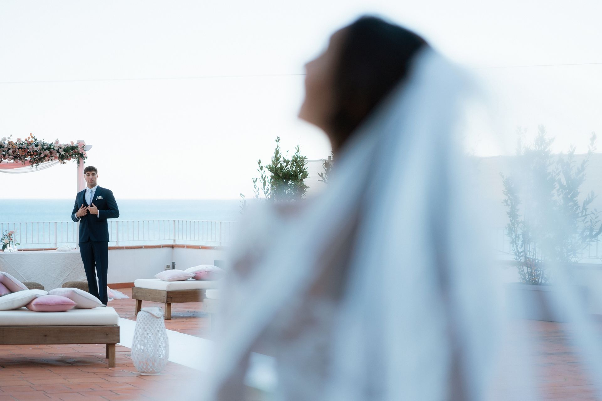 La sposa in primo piano, sfocata, con il velo, di fronte allo sposo che è in piedi davanti a un altare con vista sul mare e una terrazza decorata.