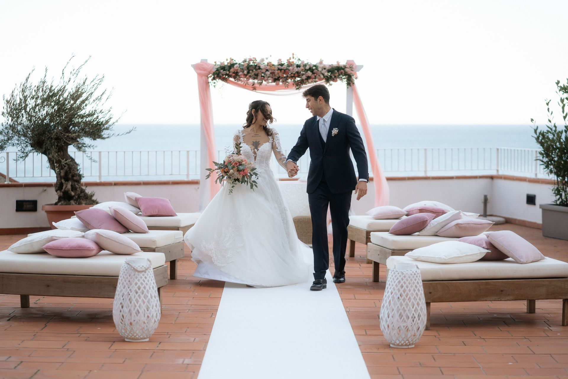 Gli sposi si tengono per mano dopo la cerimonia nuziale su una terrazza con vista sull'oceano. Percorrono la navata, circondati da sedie e decorazioni floreali rosa.