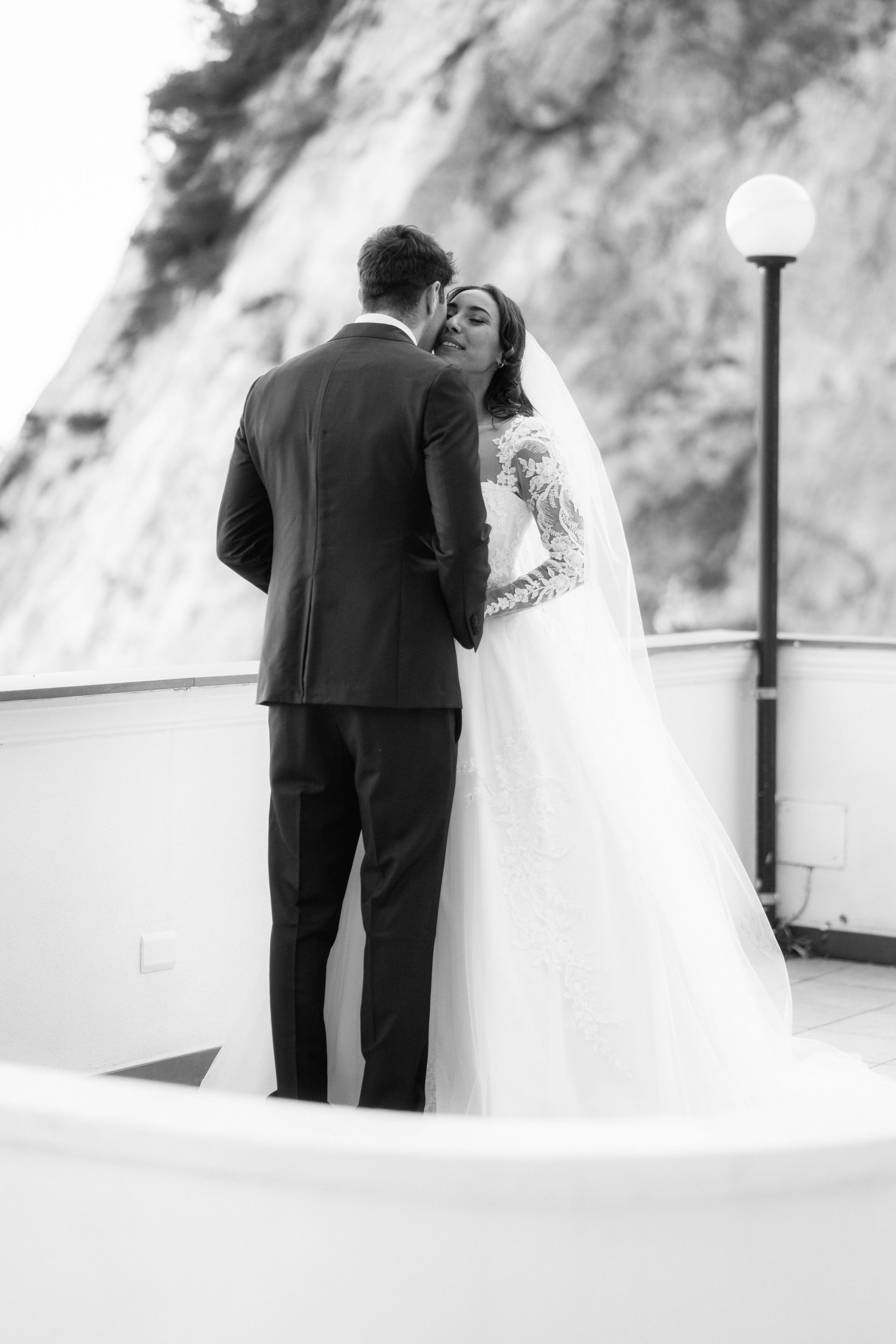 Sposi che si abbracciano su un balcone con vista su una scogliera. Foto in bianco e nero. La sposa indossa un lungo abito bianco, lo sposo un abito elegante.