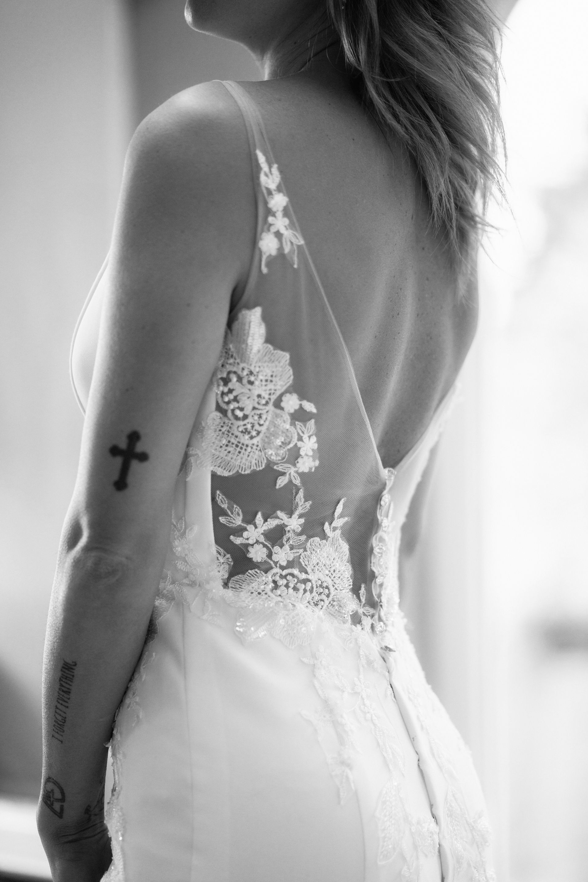 Sposa in abito da sposa, vista di spalle, con dettagli in pizzo. Ha un tatuaggio di una croce sul braccio, in piedi in un interno.