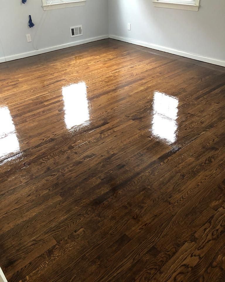 Reflective, newly-stained hardwood floor in a room with light blue walls and two windows, reflecting sunlight.