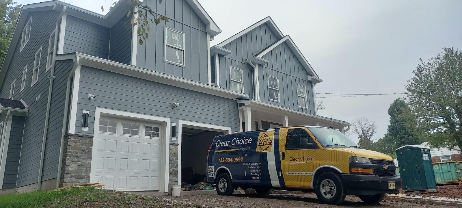 A two-story blue house with a yellow service truck parked in the driveway. A portable toilet is nearby.
