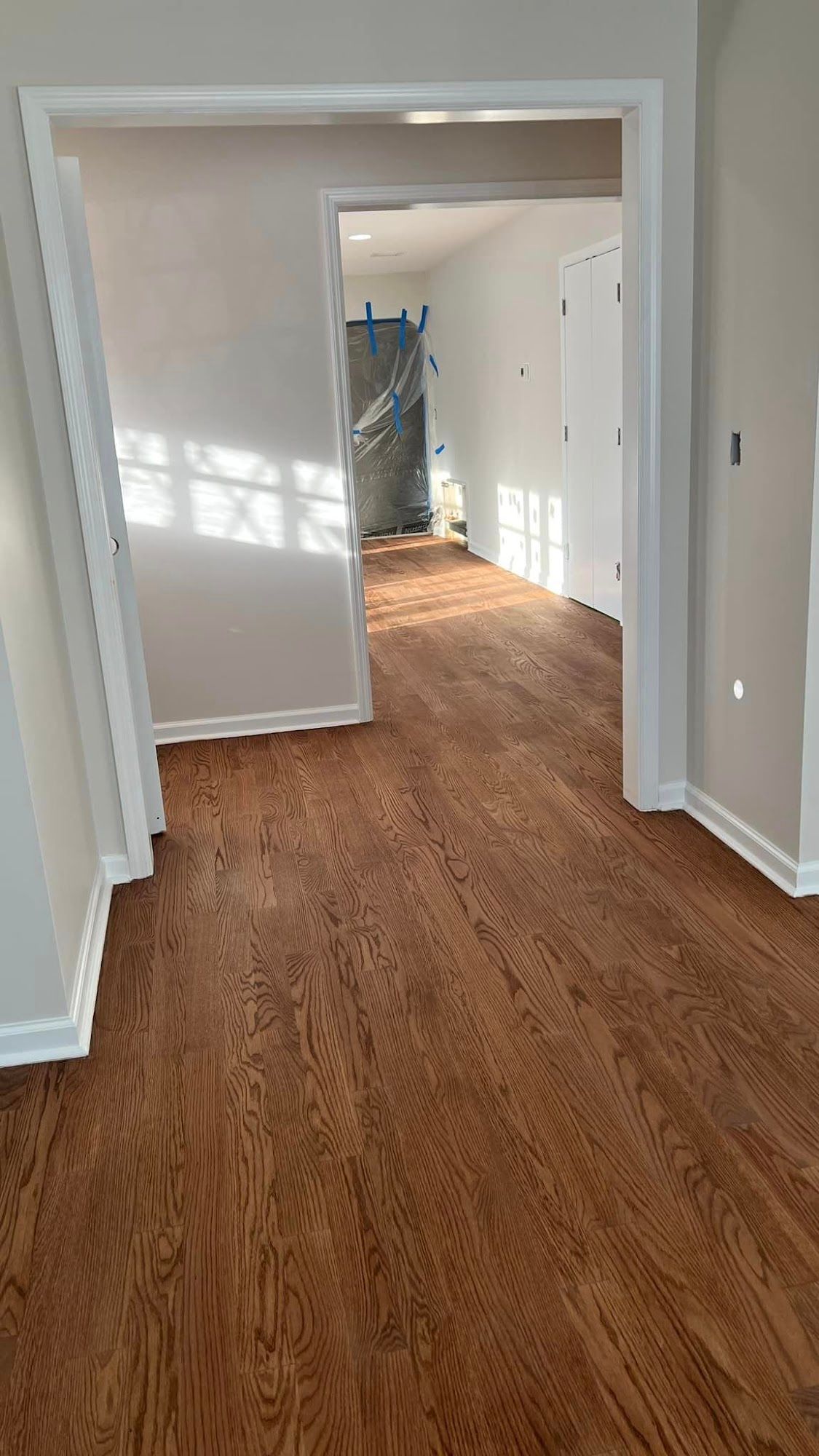 Wood floor with white walls and trim, leading to another room. Sun shines through windows.
