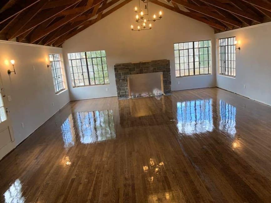 Large, empty room with high, wood-beamed ceiling, shiny wood floor, fireplace, windows, and chandelier.