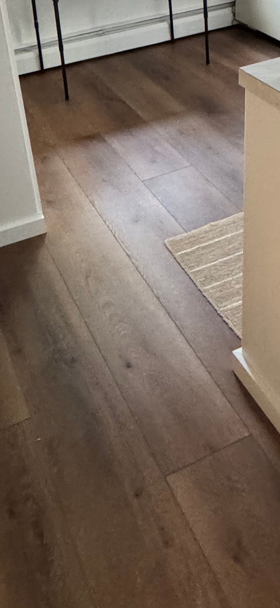 Brown wooden floor, partially covered by a neutral-toned rug, with a glimpse of a table and a wall.