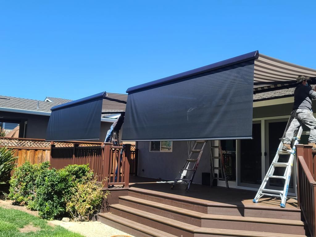 A man on a ladder is installing a black awning on a deck.