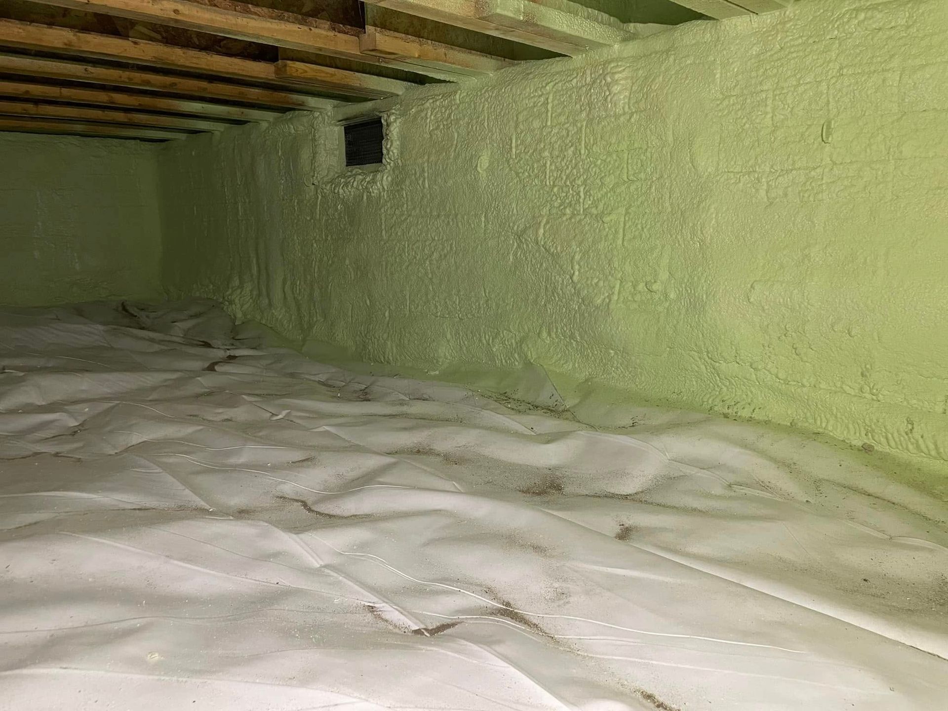 Basement crawl space with white insulation on the floor and a green painted wall under exposed beams