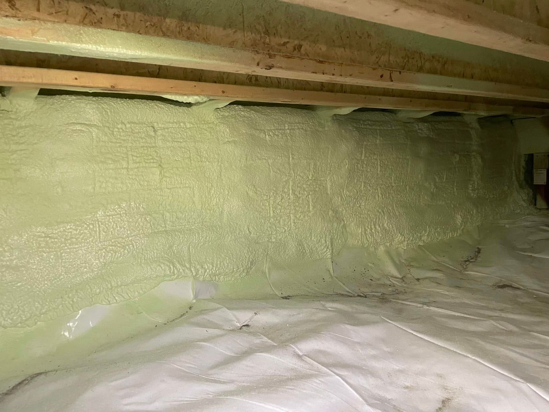Crawlspace with green spray-foam insulated wall and exposed wooden joists overhead