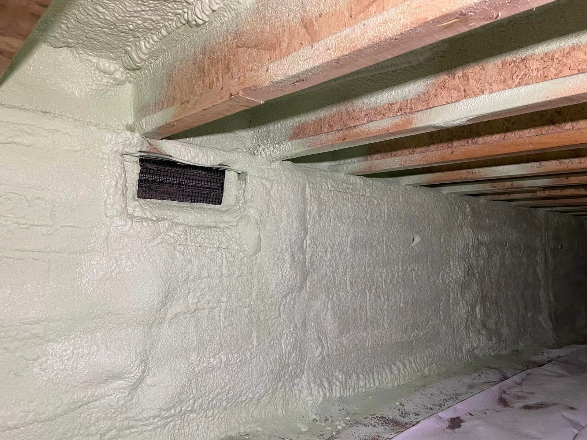 Spray-foamed basement wall with exposed ceiling beams and a small vent opening