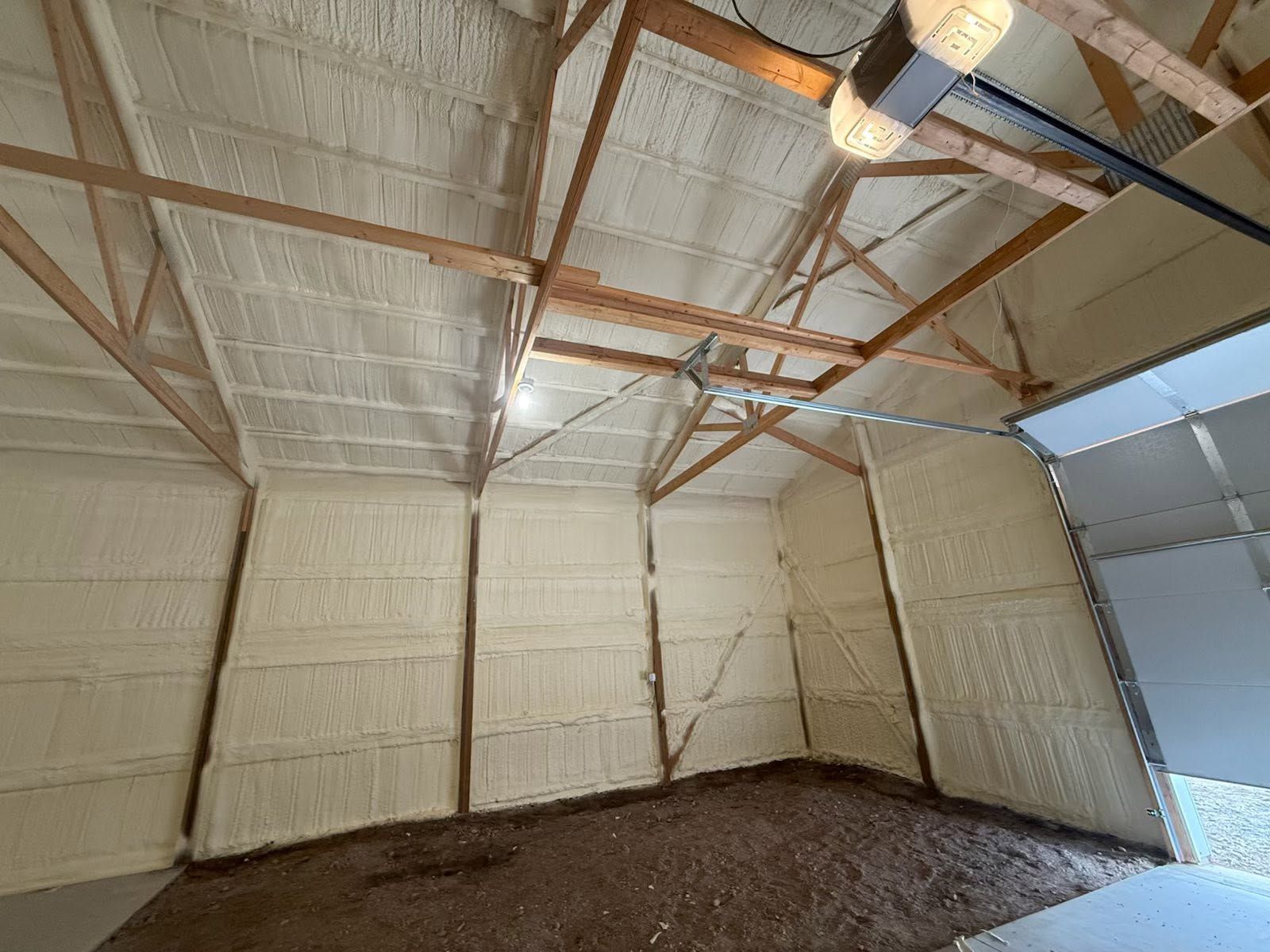 Empty insulated barn with exposed wooden rafters and a dirt floor, lit by daylight through an open side.