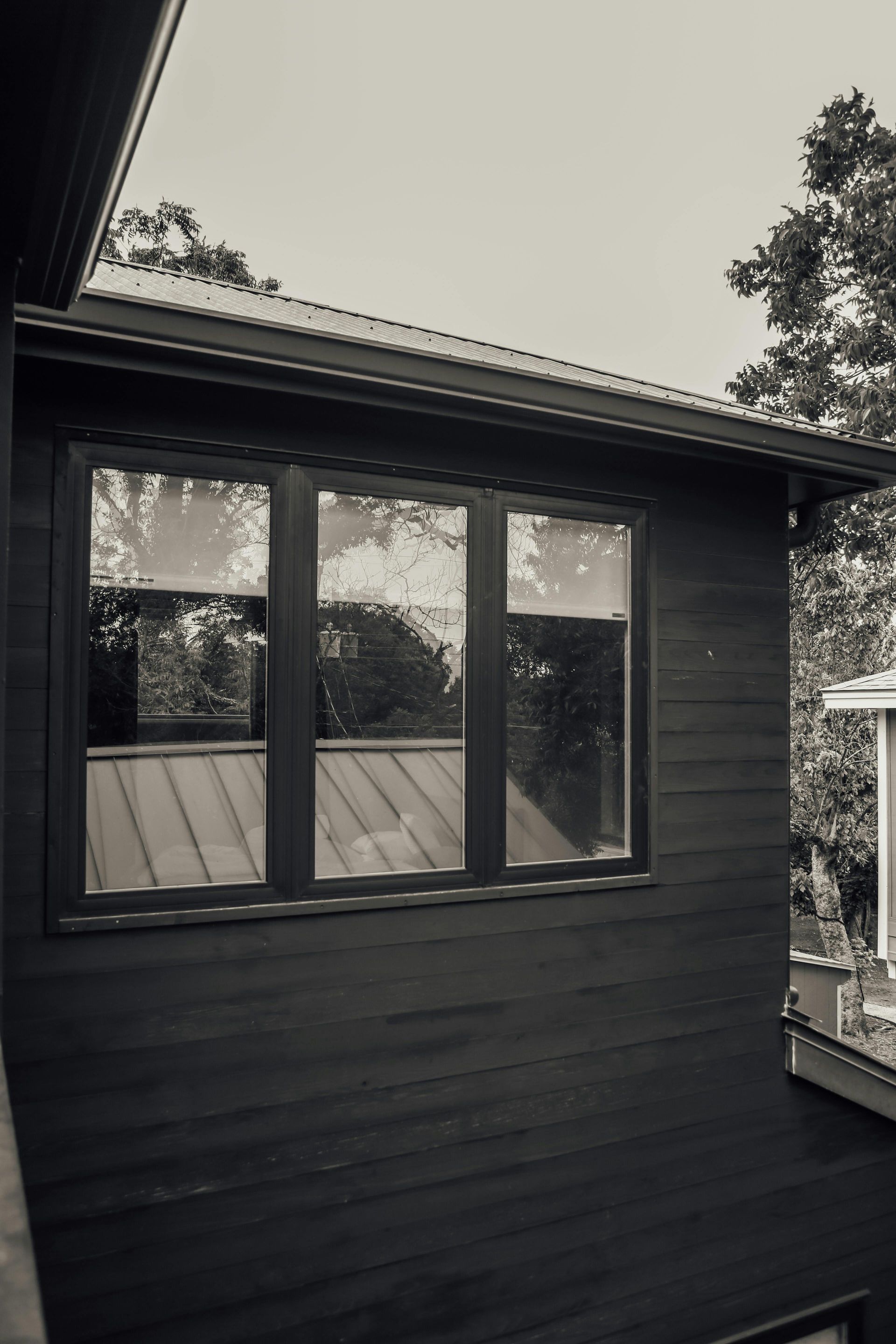 Black exterior wall with three windows under a sloped roof, trees reflected in the glass.