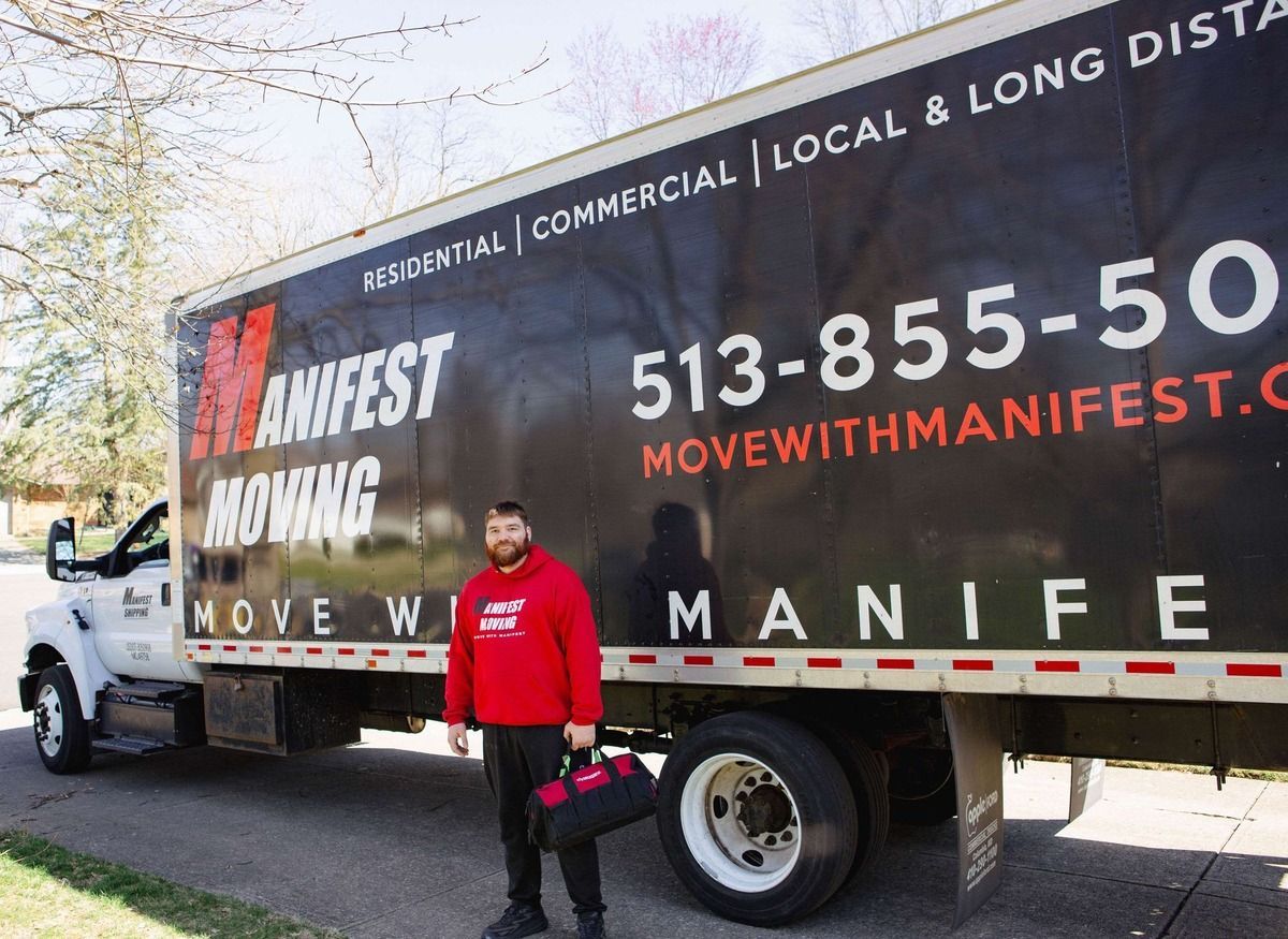 Man in red sweatshirt holding toolbox stands next to a Manifest Moving truck on a street.