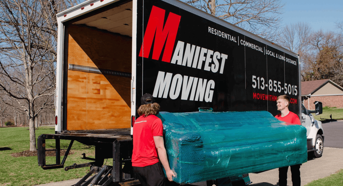 Two people in red shirts carrying a large, teal-wrapped couch toward a moving truck. Truck says 