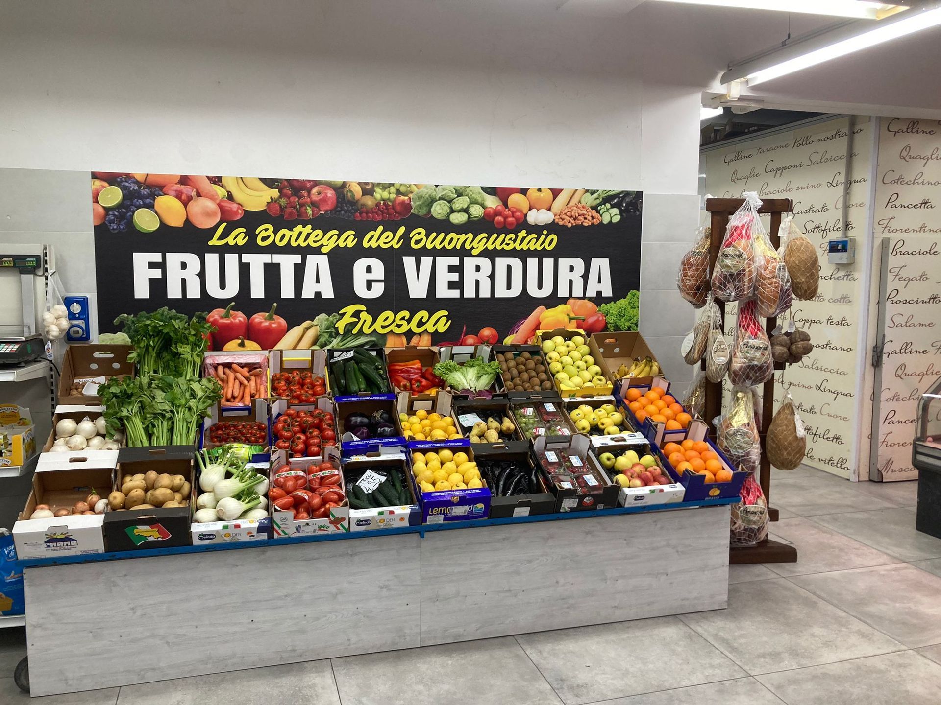 All'interno di un negozio, un'esposizione di prodotti ortofrutticoli presenta cassette di frutta e verdura fresca sotto un cartello con la scritta