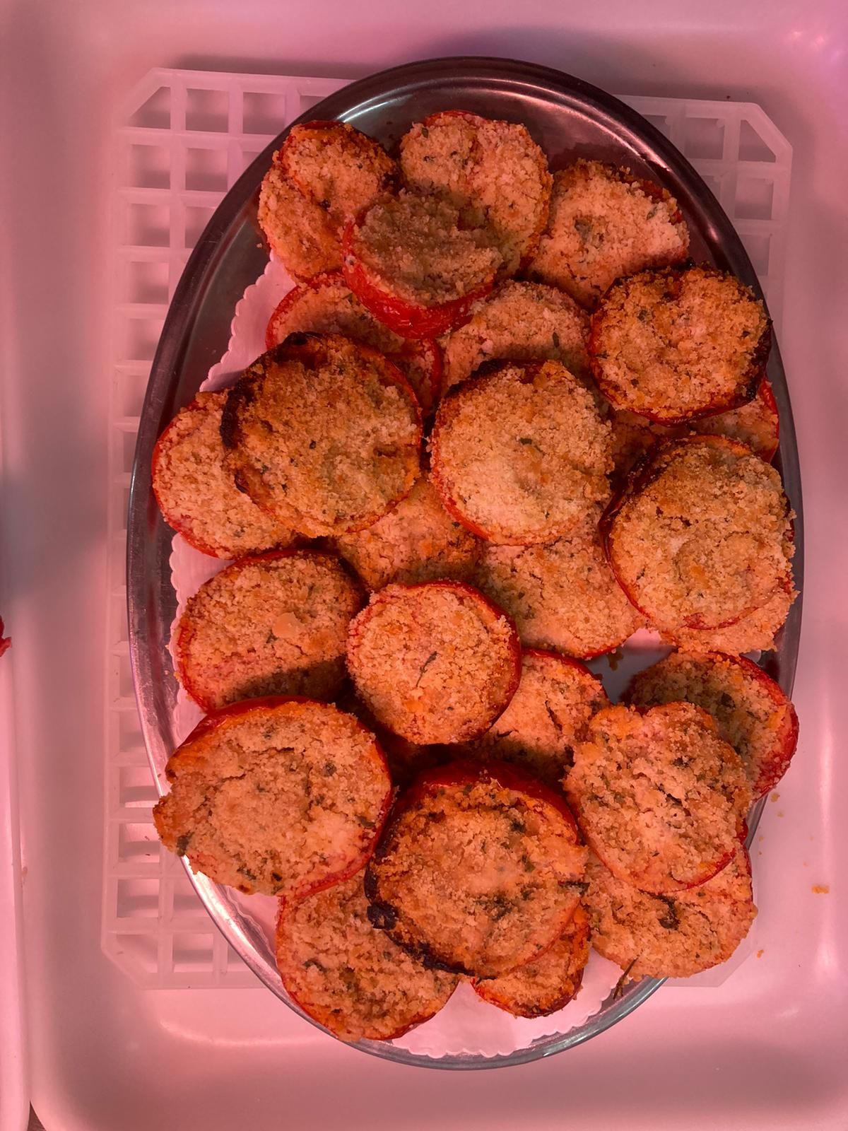 Un vassoio ovale d'argento, riempito con mezze pomodori ripiene rotonde di colore marrone dorato, è adagiato su una superficie bianca.