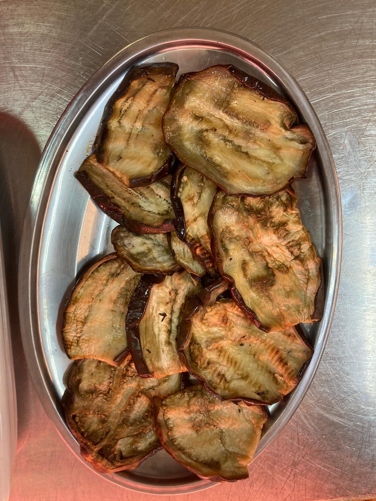 Un vassoio ovale d'argento colmo di melanzane grigliate a fette, dorate e croccanti, disposte su una superficie metallica.