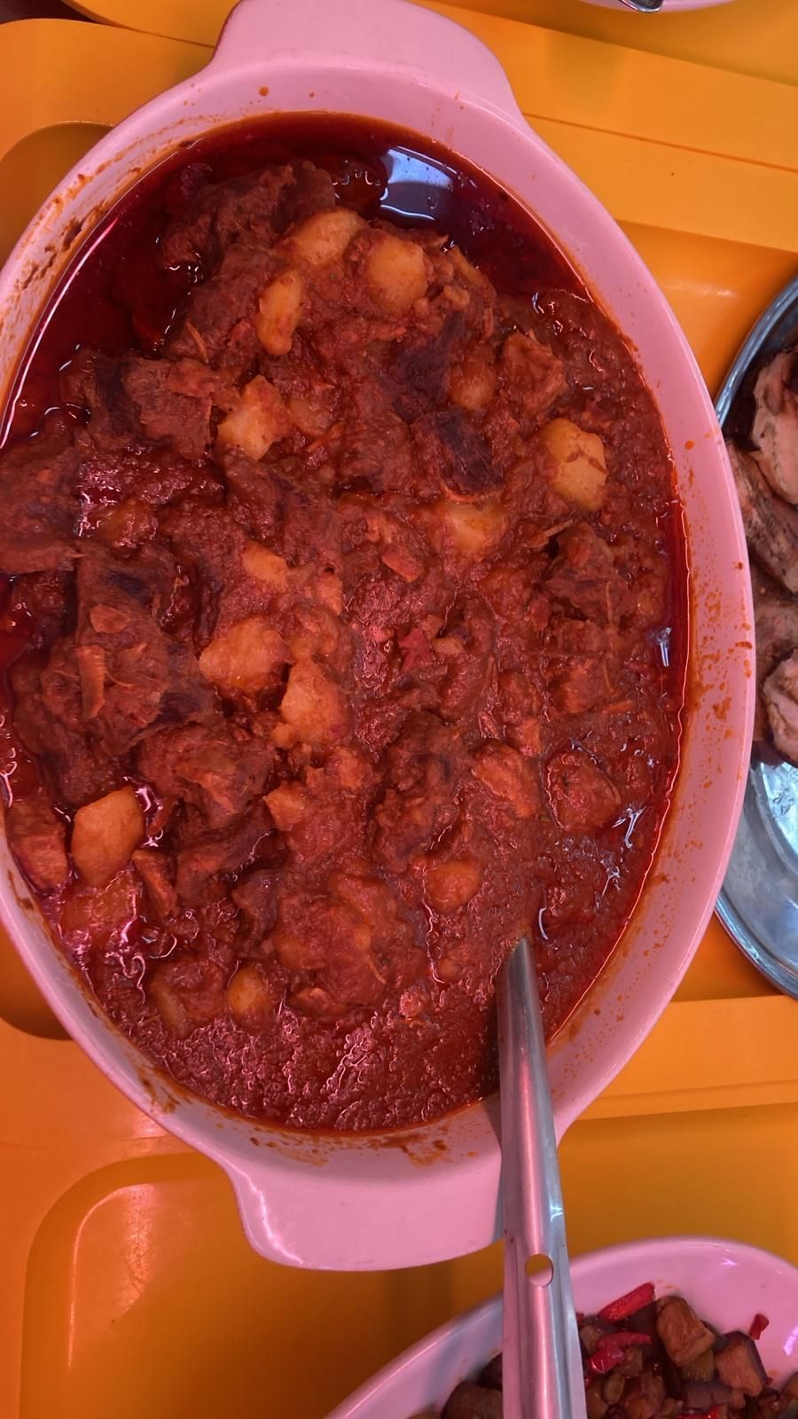 Una ciotola ovale riempita con uno stufato di carne e patate di colore marrone rossastro scuro, adagiata su un vassoio giallo brillante.