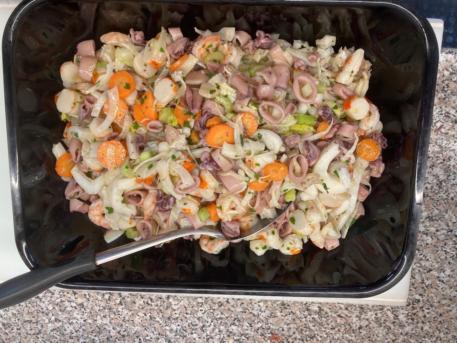 Un contenitore nero contiene un'insalata di calamari a fette, carote arancioni a rondelle e sedano verde, accompagnata da un cucchiaio di metallo.