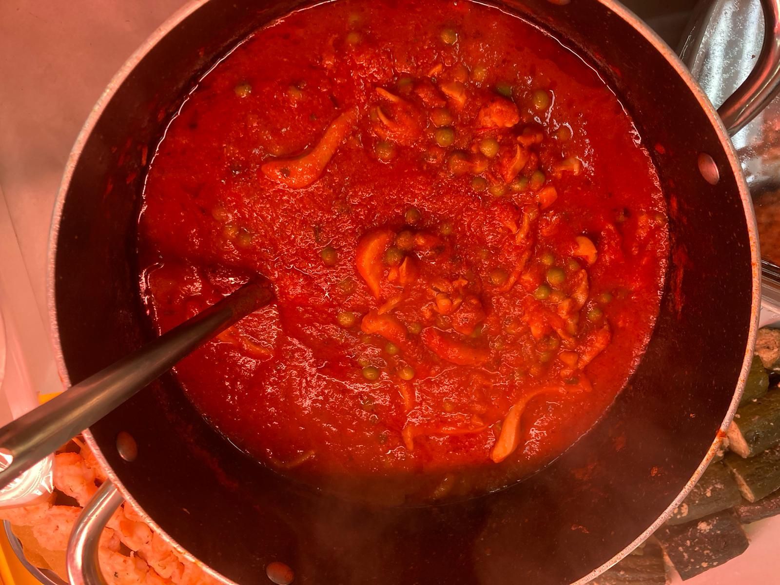 Una grande pentola di metallo contenente uno stufato a base di pomodoro con piselli e straccetti di carne, con un cucchiaio da portata appoggiato all'interno.