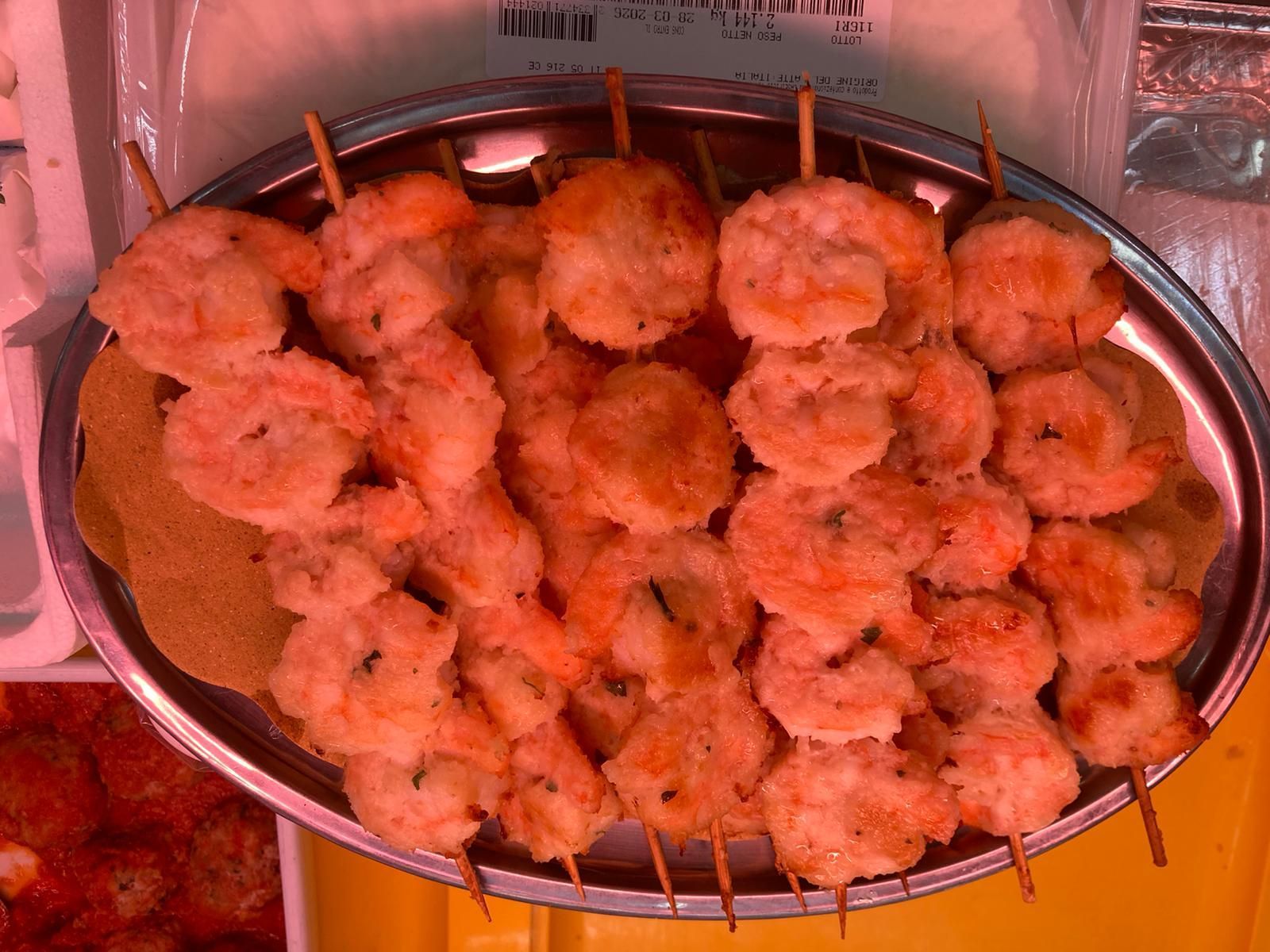 Su un vassoio d'argento sono disposti diversi spiedini di legno contenenti gamberi cotti di colore rosa, adagiati su un foglio di pergamena marrone chiaro.