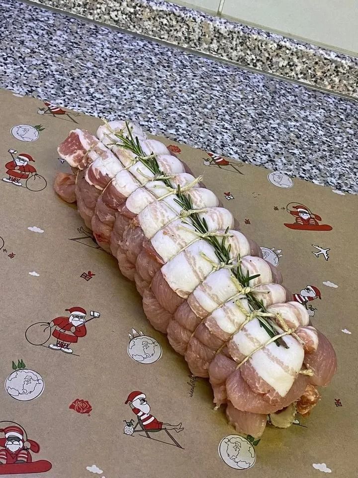 Un arrosto di maiale crudo, legato e condito con rosmarino, adagiato su carta da regalo marrone a tema natalizio con motivi di Babbo Natale.