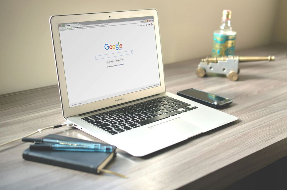 A silver laptop displaying the Google homepage sits on a desk next to a notebook, pens, a smartphone, and a small cannon.