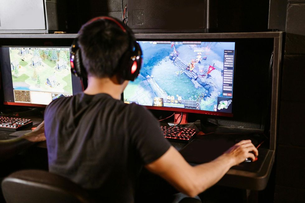 A person in a black t-shirt and gaming headset plays a video game on a dual-monitor computer setup in a dark room.