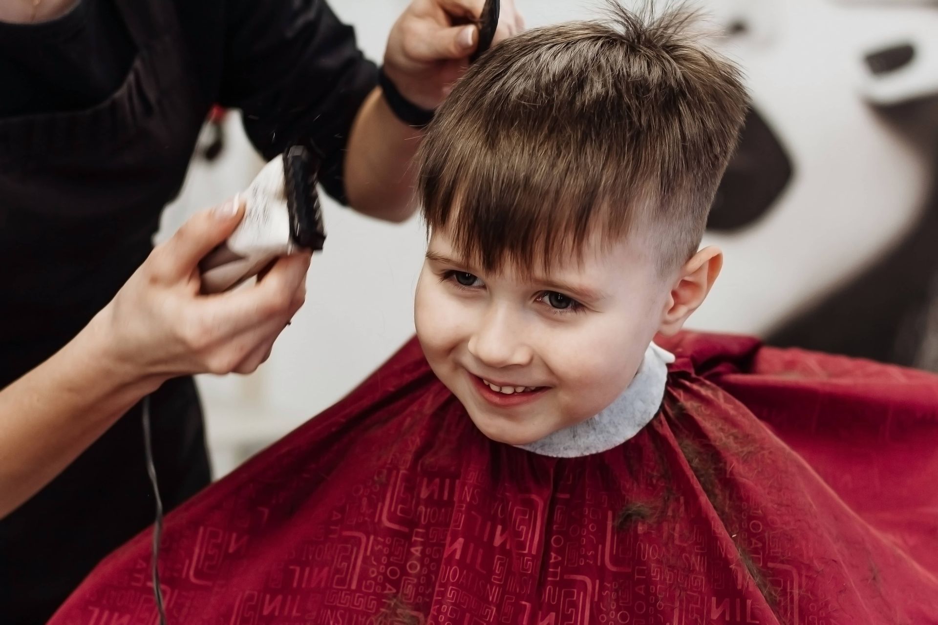 Boy smiles while getting a haircut with a clipper in a salon.
