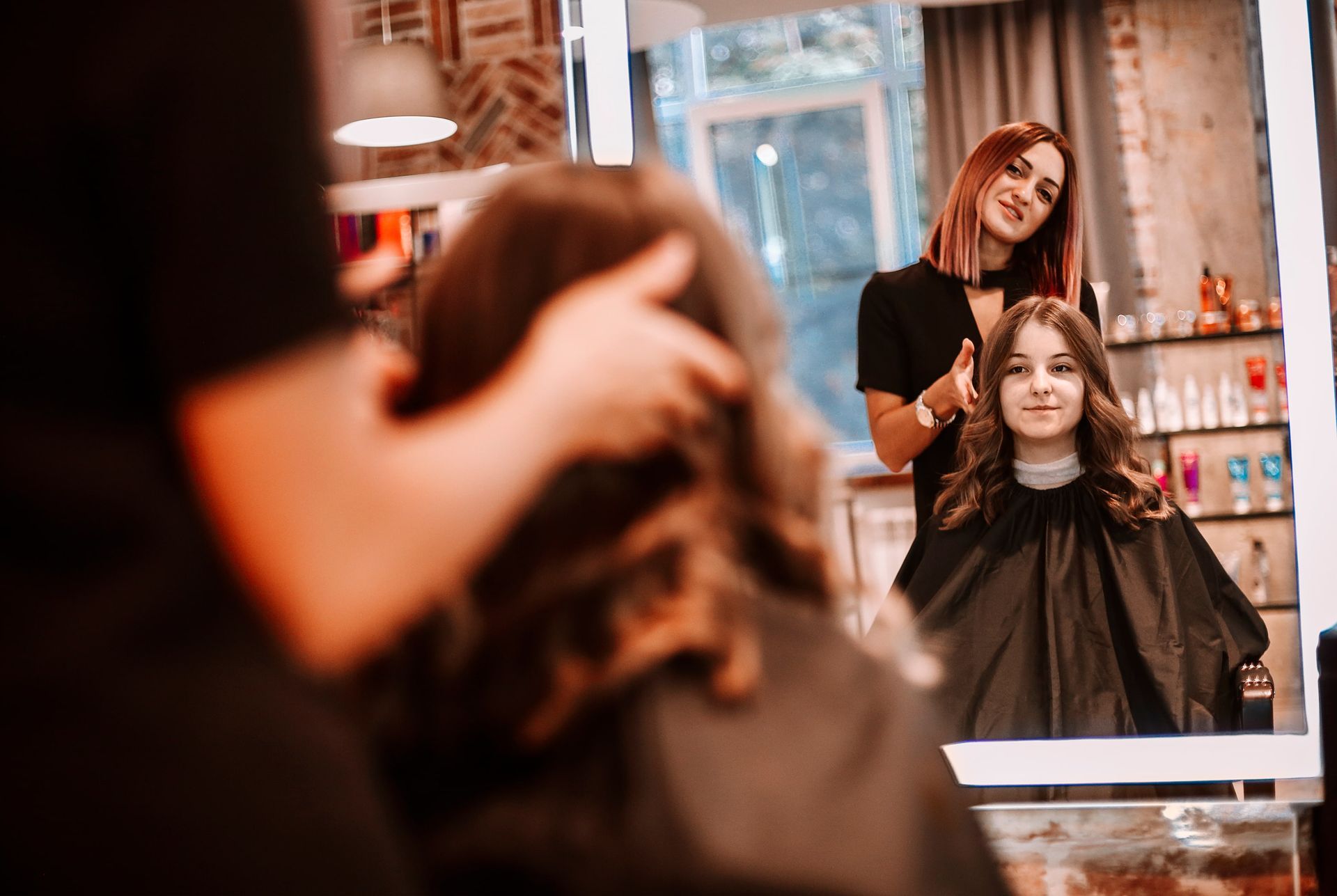Hair stylist with a customer in a salon; mirror reflection shows the stylist adjusting the client's wavy hair.