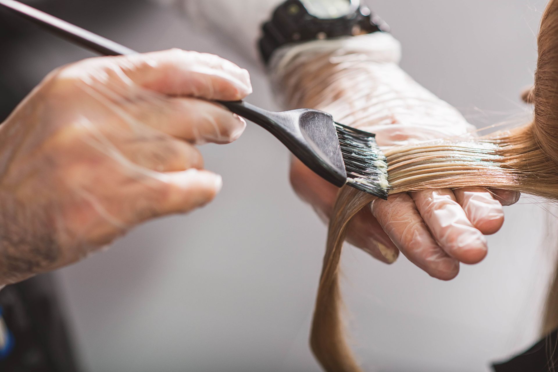 Hands in gloves applying hair dye with a brush to blonde hair.