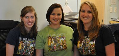 Three women are posing for a picture and one of them is wearing a shirt that says all are welcome