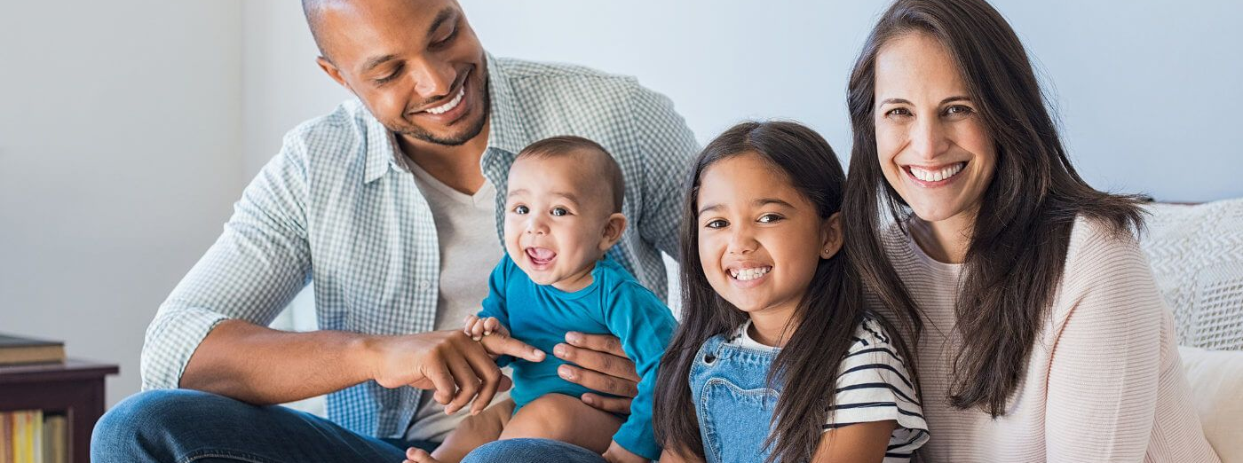 Family of four smiling, sitting together. Baby held by father. Two parents and two children on a couch.